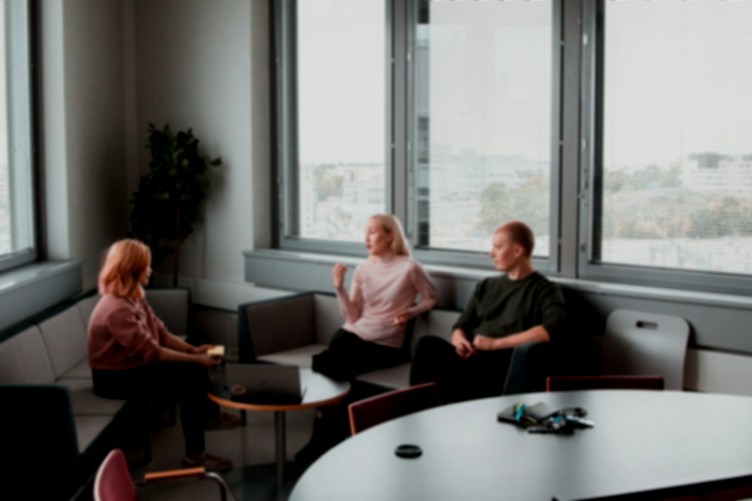 Three people sitting in a circle in a room with large windows, engaged in conversation. One woman with red hair, another woman with blonde hair, and a man with short hair. Furniture includes chairs and small tables.