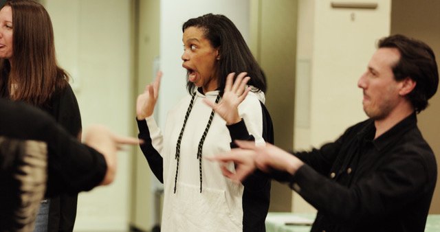 Three people in a room, with one woman in the center showing an surprised or expressive reaction as she talks, while the others listen or interact.