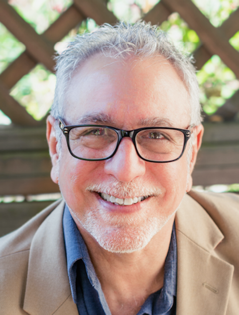 Smiling older man with gray hair and glasses, outdoors with a wooden fence and green foliage in the background.