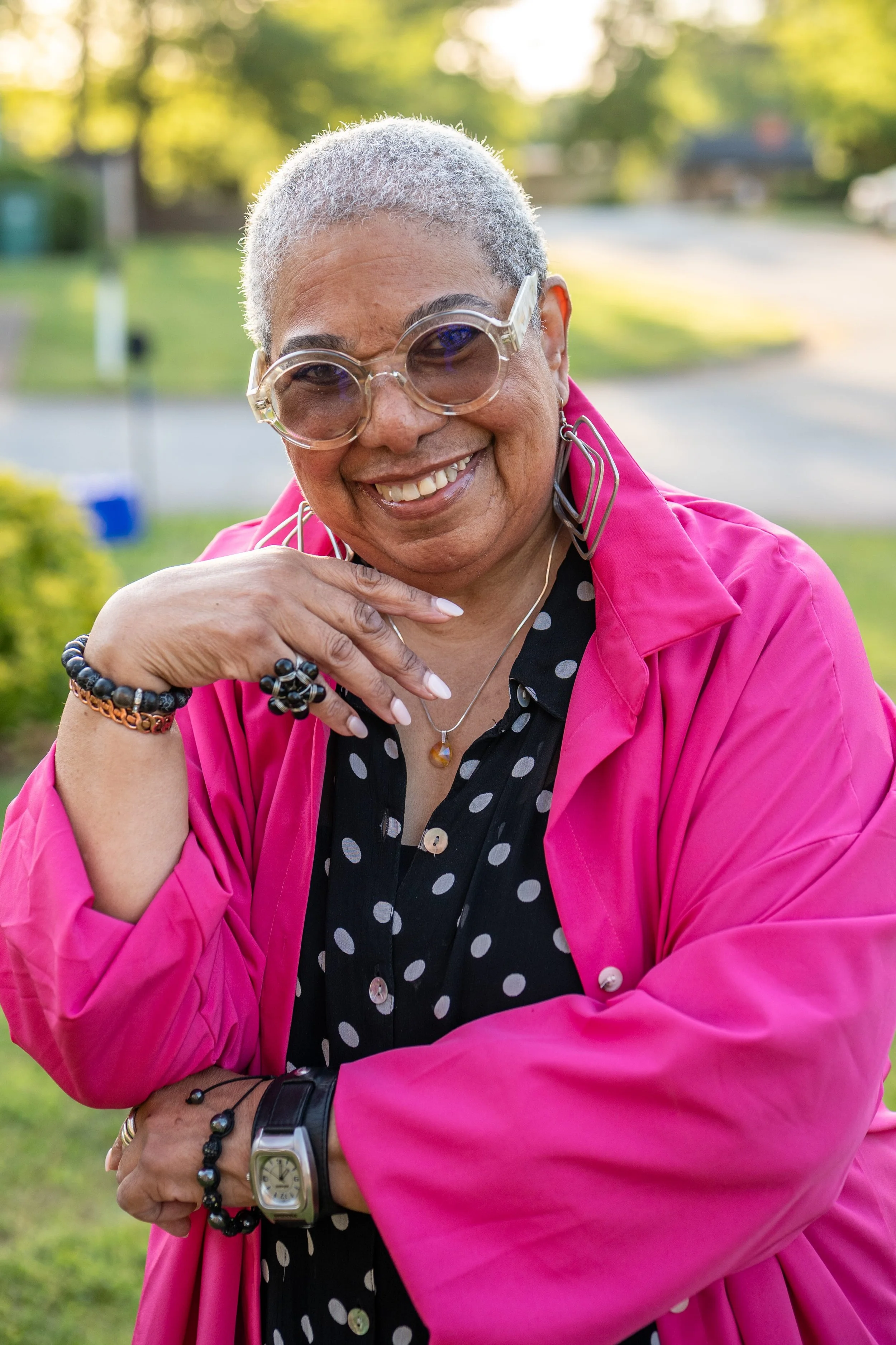 A woman with short gray hair wearing large round glasses, a pink jacket, black and white polka dot shirt, and jewelry, smiling outdoors with a green background.