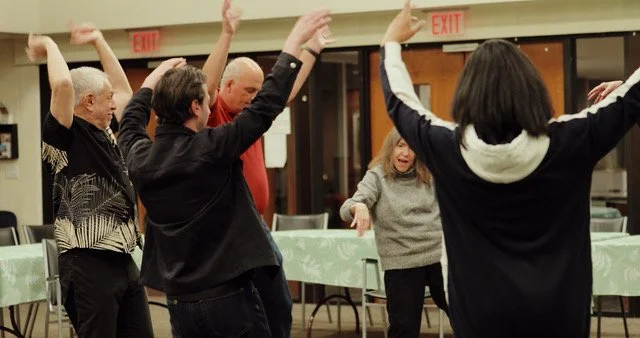 A group of people dancing and having fun in a room with tables and chairs.