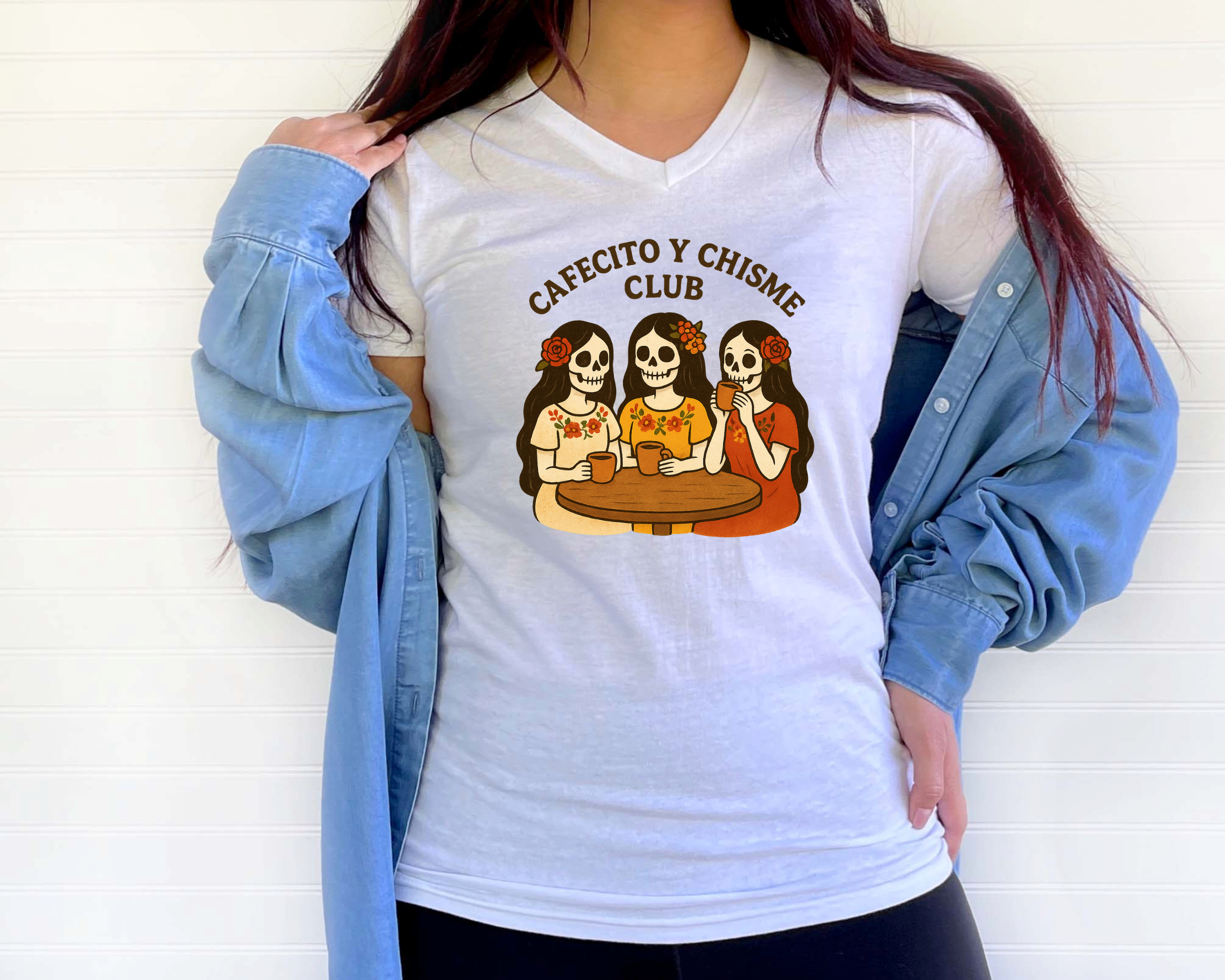A woman wears a white T-shirt with a graphic of three sugar skull women at a table, holding coffee mugs, and the text 'CafeCito y Chisme Club.' She has a blue jacket draped over her shoulders and a white background.