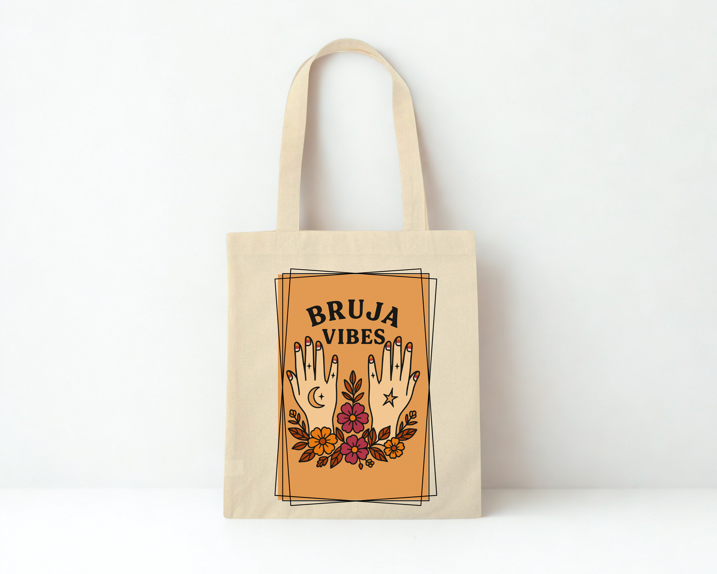A beige tote bag with a design featuring two hands with decorative tattoos, flowers, moon, and star, along with the words "BRUJA VIBES" in bold black letters.