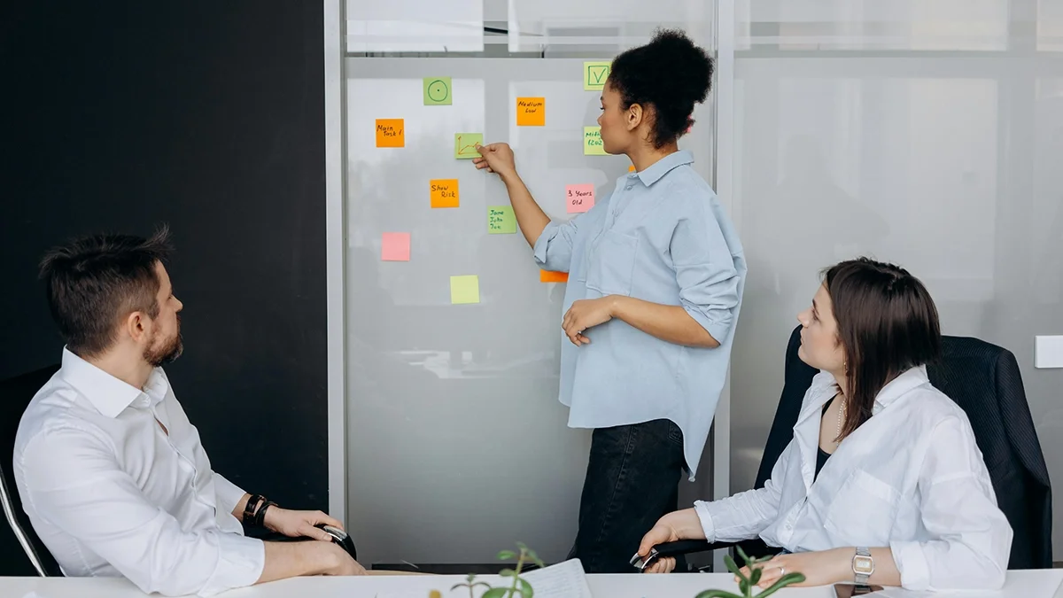 Three people in a meeting room, one woman presenting with sticky notes on a whiteboard, two others listening at the table.