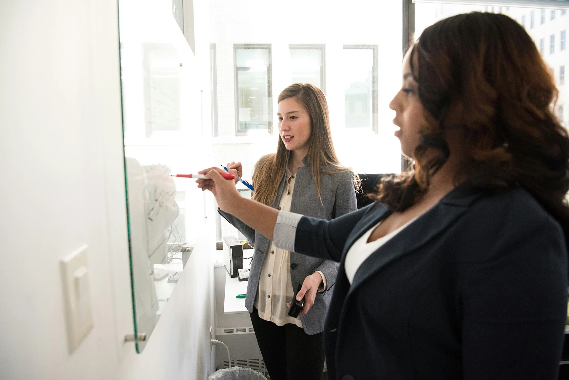 Two women in professional attire writing on a glass board in an office.