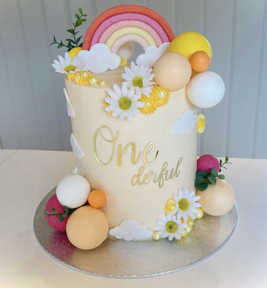 A birthday cake decorated with colorful balloons, daisies, clouds, a rainbow, and greenery. The cake has the words "One derful" written in gold lettering on the front.