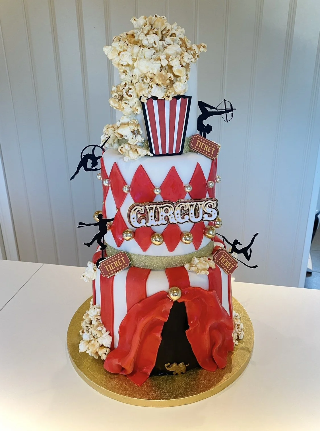 A circus-themed cake with two tiers decorated in red, white, and gold, featuring popcorn, silhouettes of acrobats, tickets, and a clown's mouth entrance.
