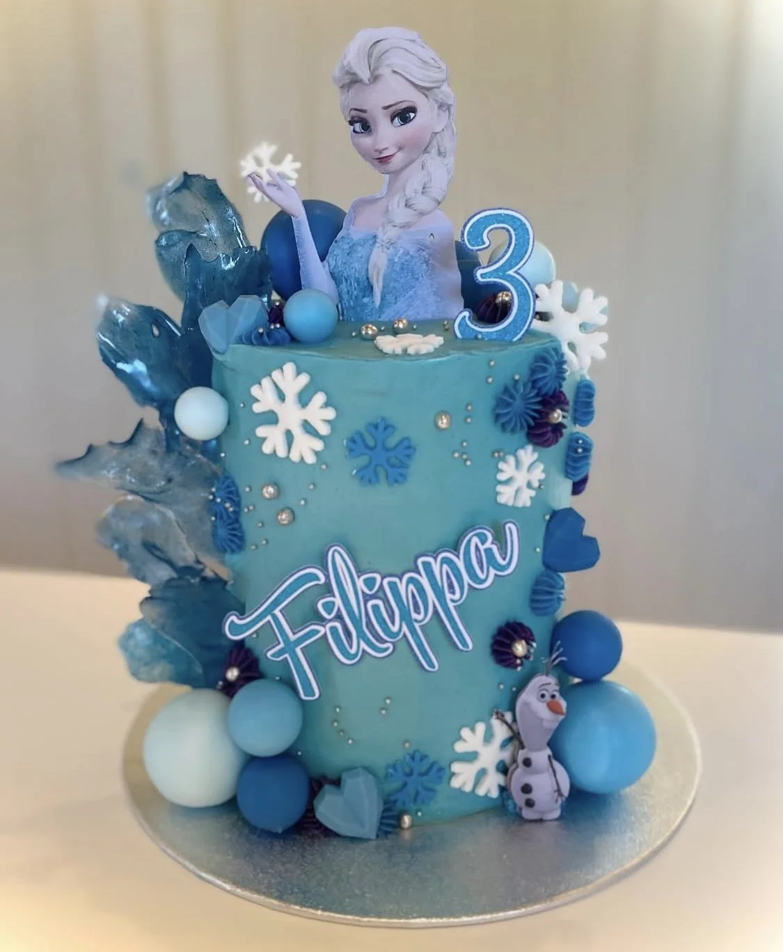A blue birthday cake with an Elsa figure from Frozen on top, snowflake decorations, and the name 'Filippao' written on it. The cake is decorated with blue and white balls, snowflake and icy decorations, and a small Olaf character at the base. There i