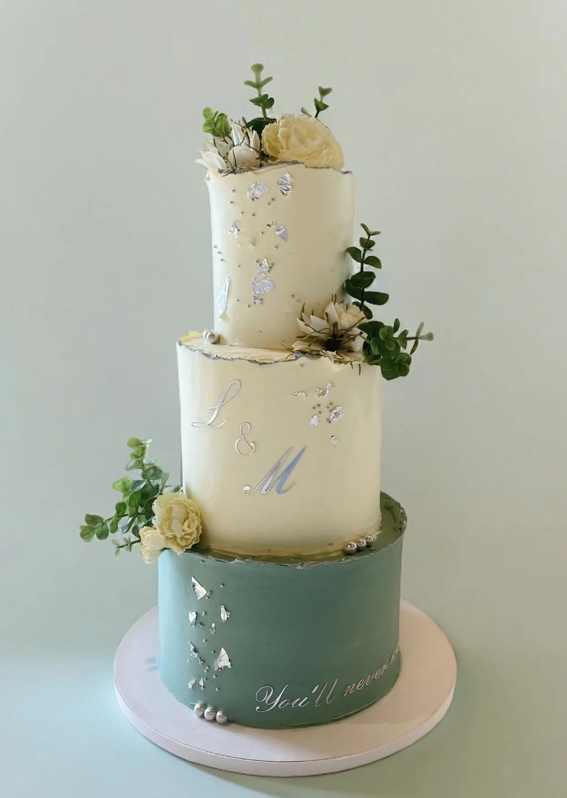 Three-tiered wedding cake with white and green icing, decorated with silver accents, flowers, and greenery, with the words "You'll never" visible on the bottom tier.