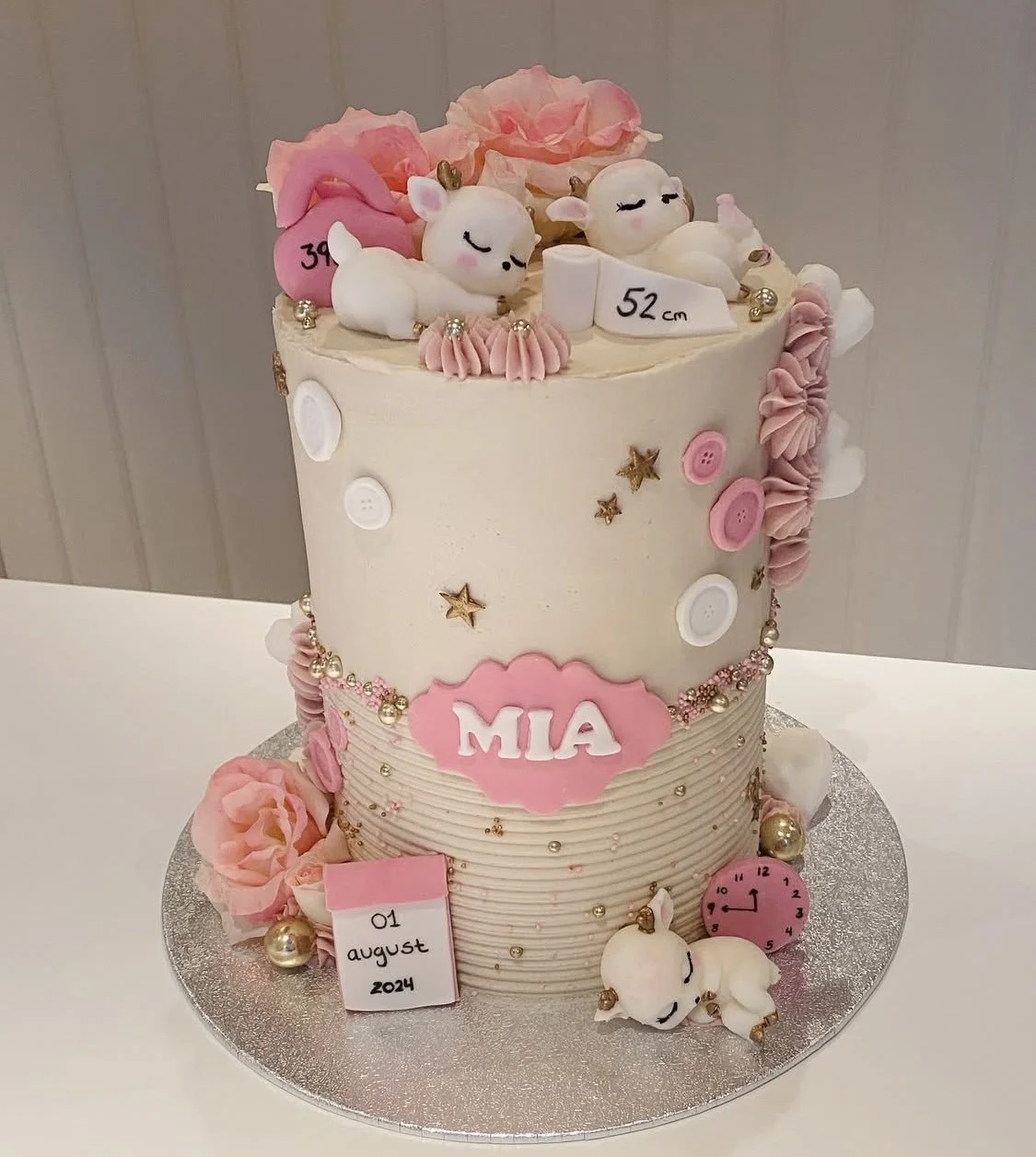 A pink and white birthday cake decorated with plush unicorns, pink and white flowers, buttons, stars, and beads, with a pink plaque reading 'MIA,' indicating a celebration on August 1, 2024.