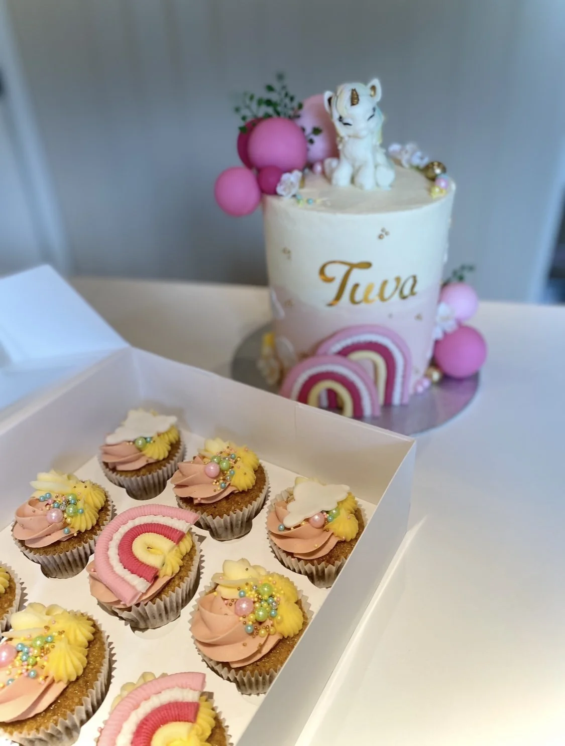 Colorful cupcakes with pink, yellow, and white frosting, decorated with sprinkles, arranged in a box in front of a pastel birthday cake with a unicorn figurine and pink, purple, and gold decorations.