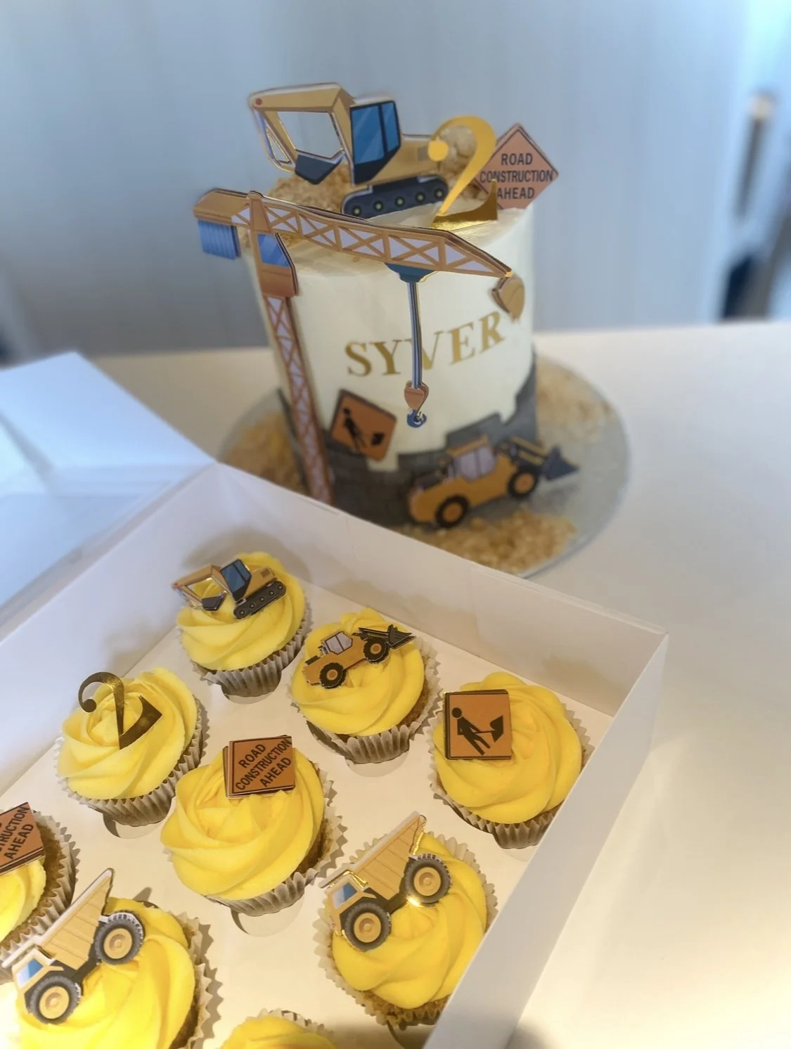 Construction-themed cupcakes with yellow frosting and construction vehicle toppers, set on a white tray, with a larger cake in the background decorated with construction signs, a crane, and a building theme.