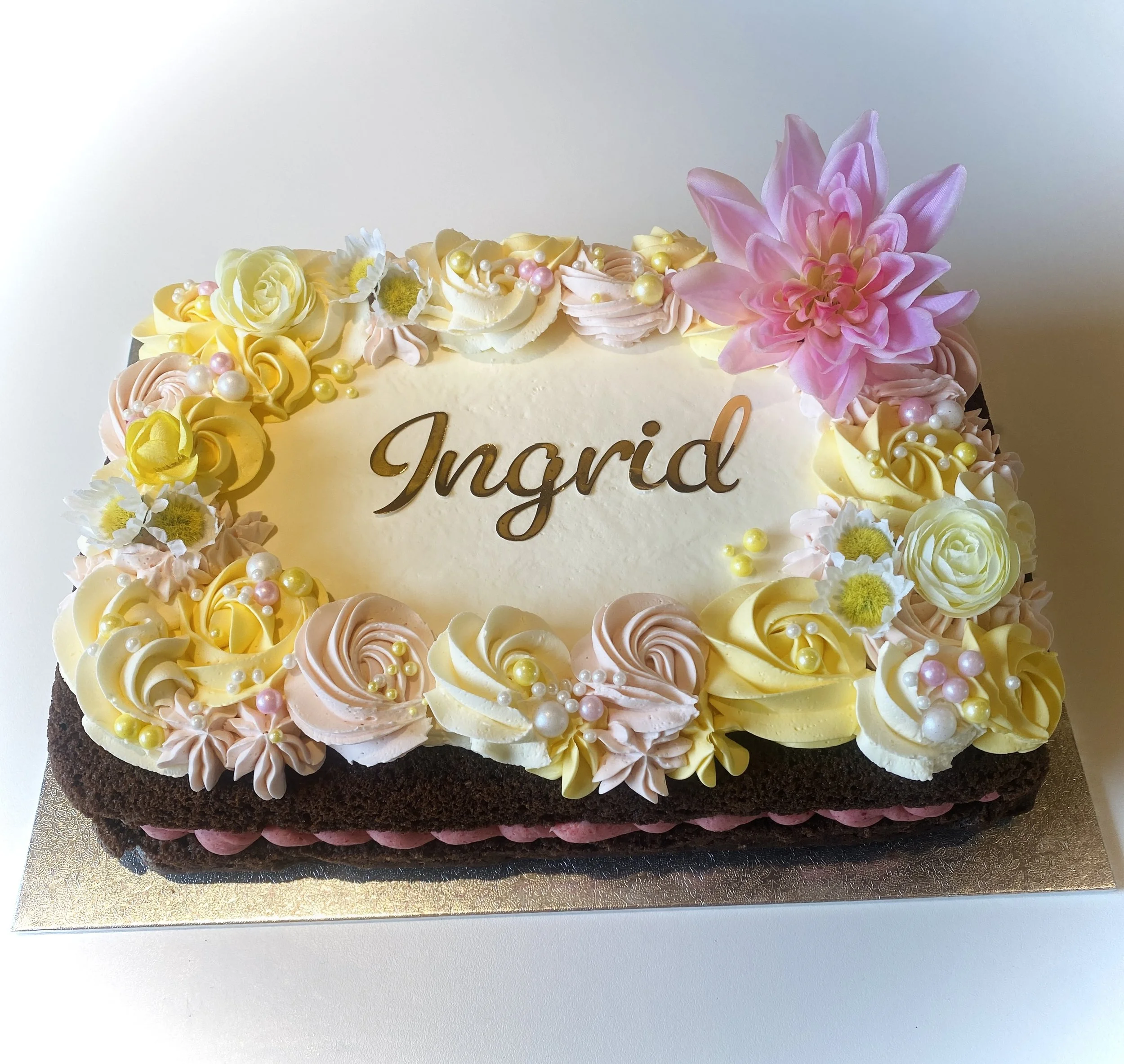 Decorative birthday cake with the name Ingrid written on top, adorned with pink, white, and yellow flowers, pearls, and cream swirls, sitting on a gold cake board.