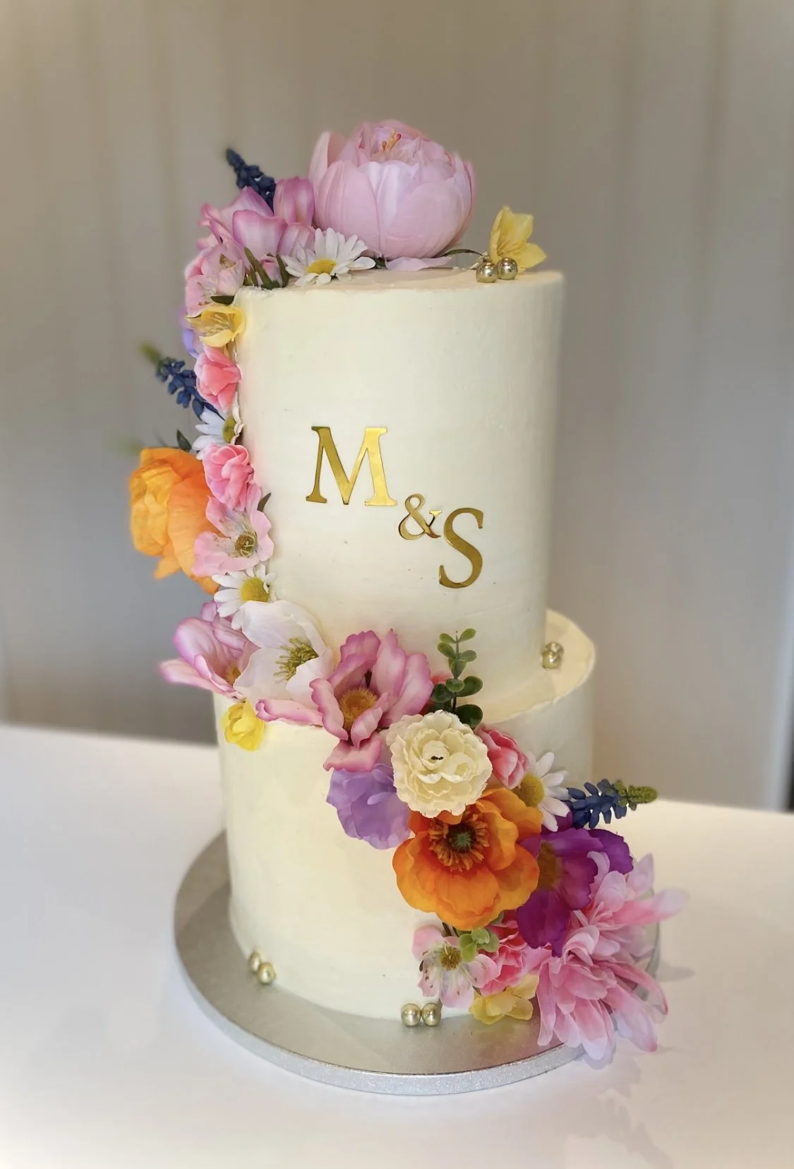 A two-tiered white wedding cake decorated with colorful flowers and gold initials 'M & S' in the center.
