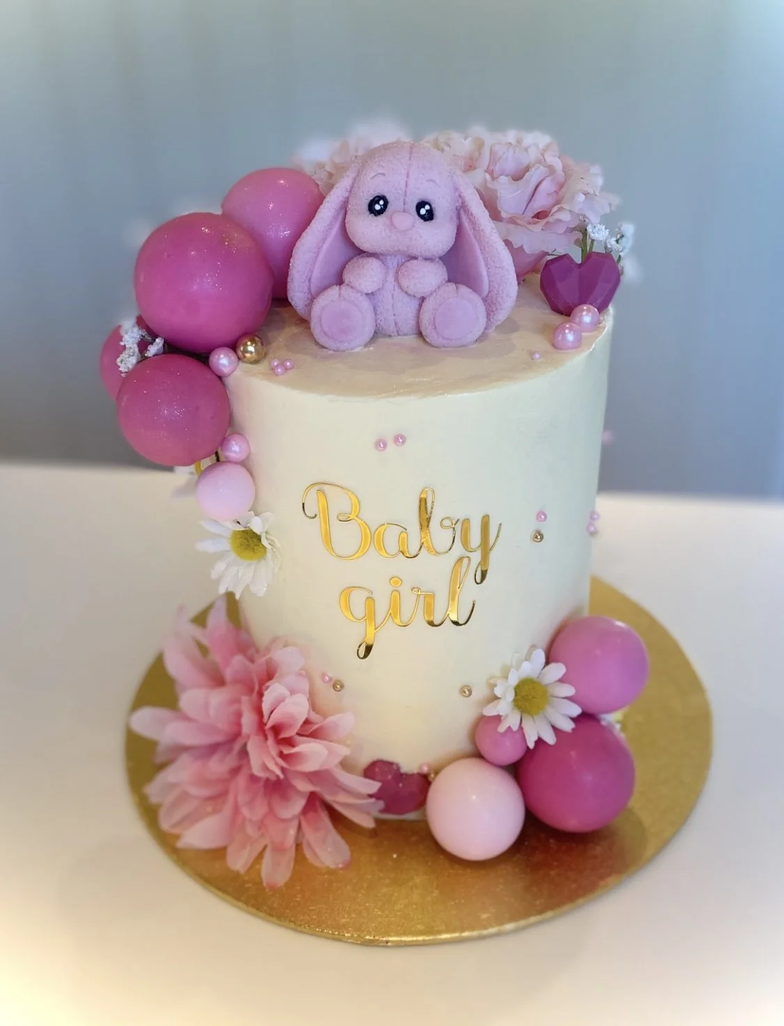 A baby girl-themed birthday cake decorated with pink flowers, pink and white balls, and a cute pink bunny plush on top. Gold text on the cake reads "Baby Girl."