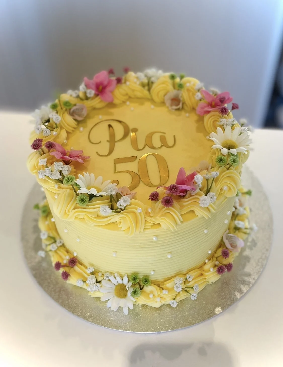 Yellow birthday cake decorated with flowers, with the words 'Pia 50' written on top.