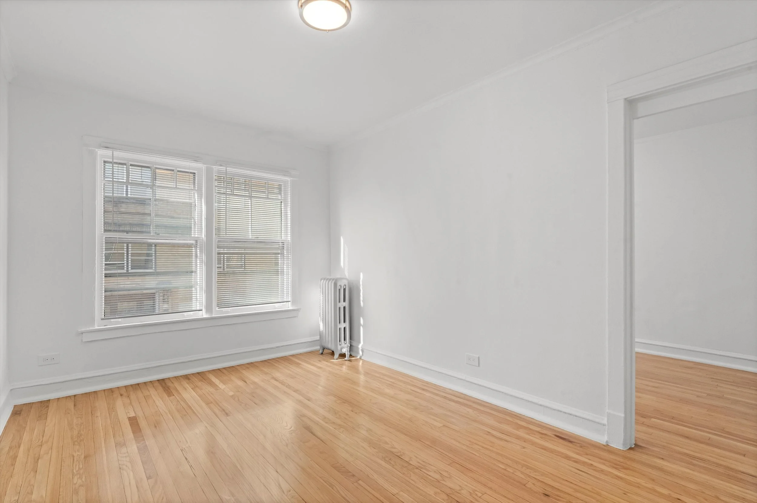 Empty room with white walls, hardwood floors, three windows with blinds, a radiator, and an open doorway leading to another room.