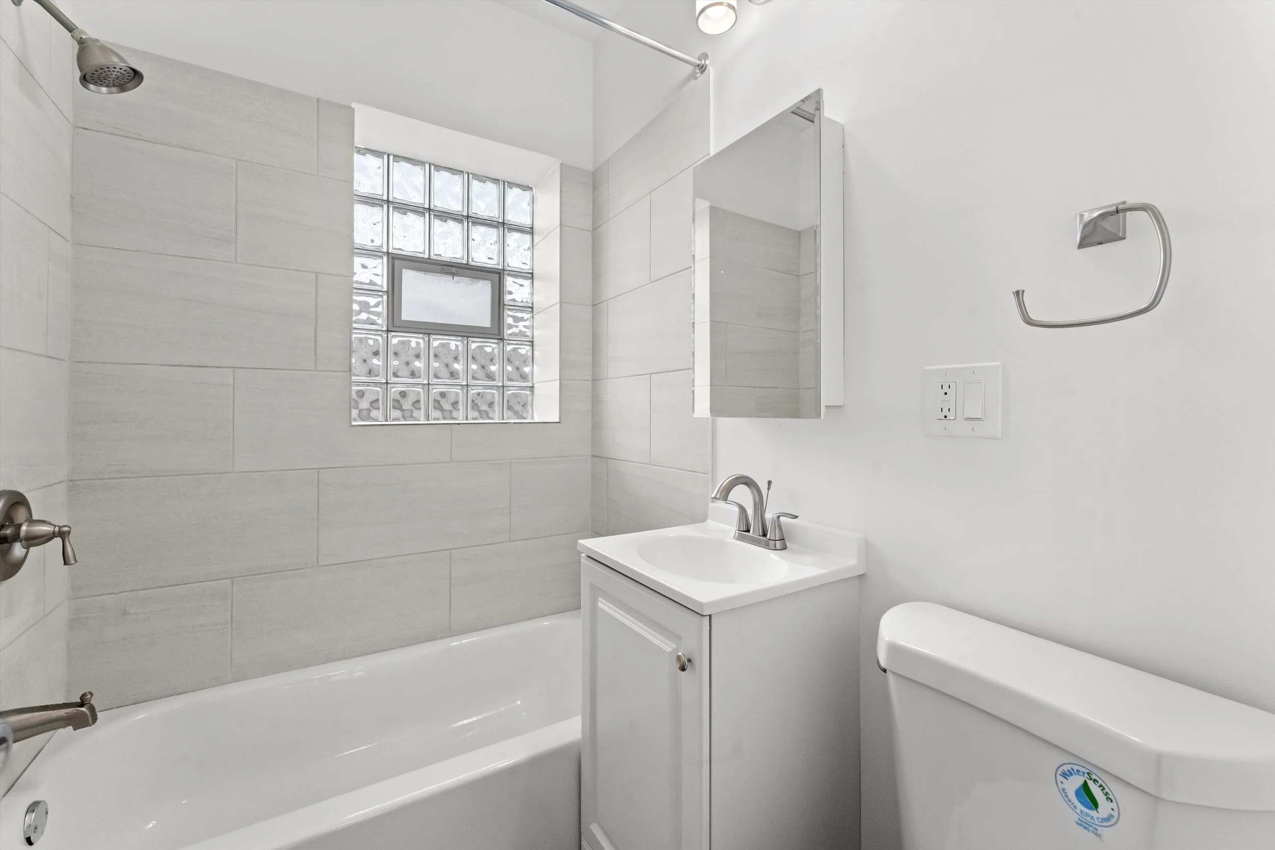 A small bathroom featuring a bathtub with a shower head, a white vanity with a sink and mirror, a toilet, and a window made of glass blocks.