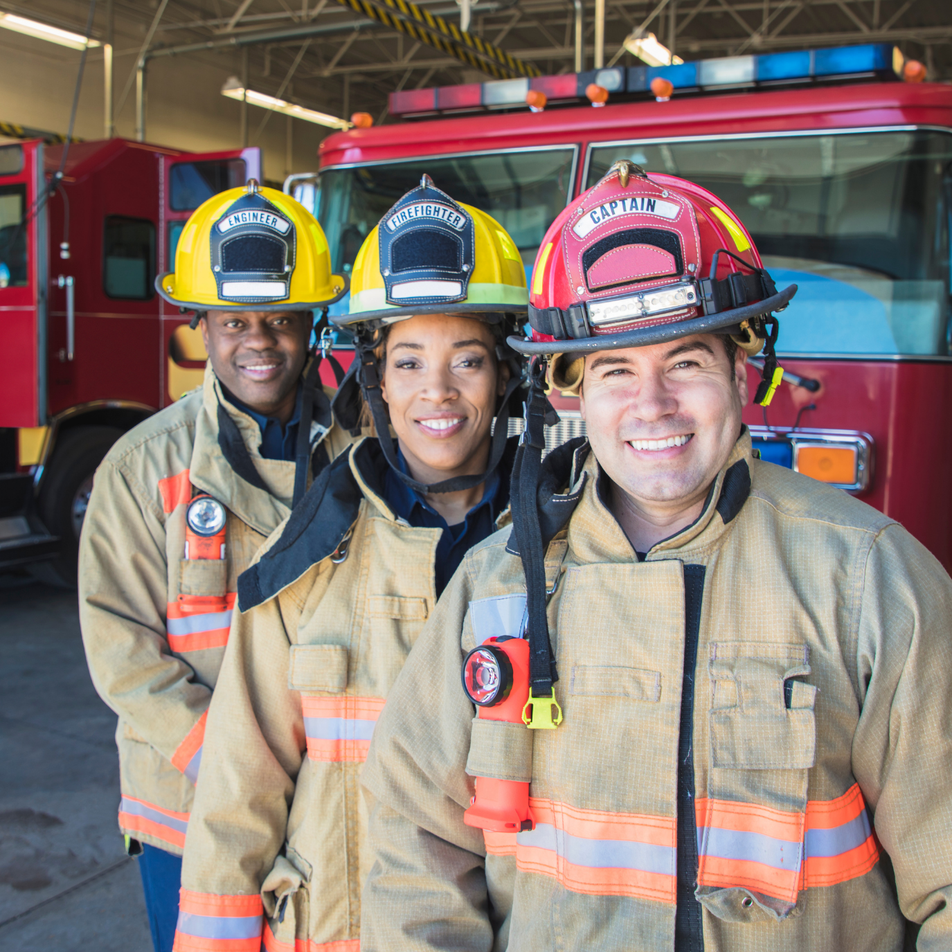 Three firefighters in firefighting gear, smiling, standing in front of a fire truck inside a fire station.