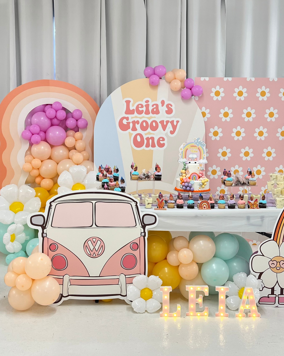 Pink and purple balloons arranged in a cluster at a pastel-colored party or event, with a pink backdrop featuring the name 'Laura' in purple letters.