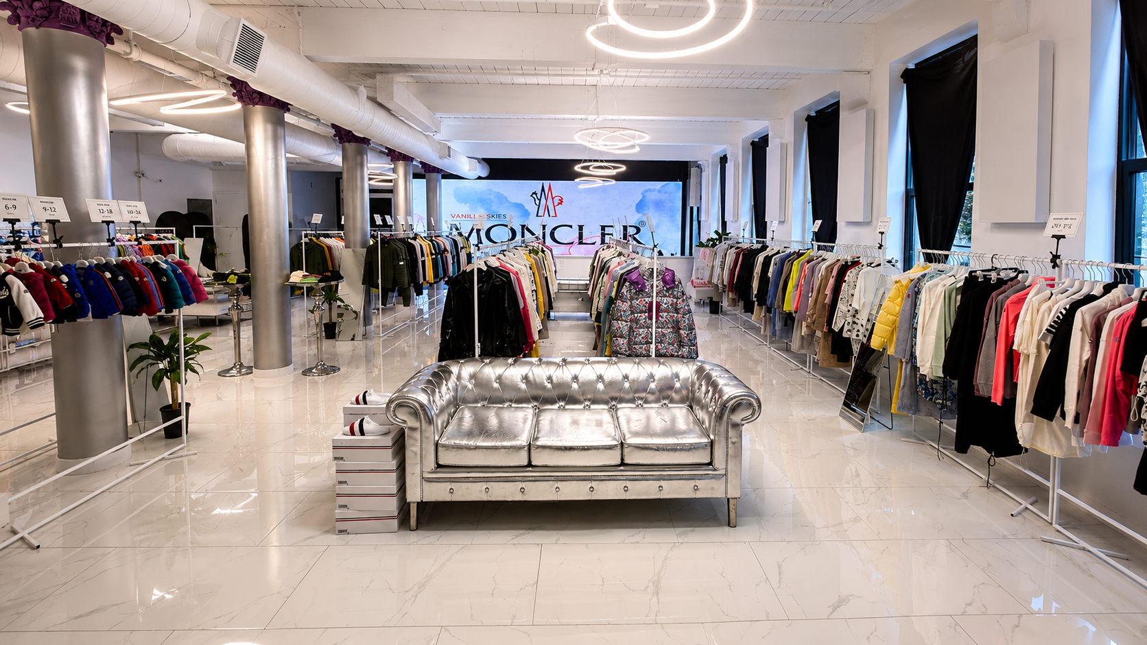 Clothing racks with various jackets and coats in a retail store, with a central palm plant and gold stanchions with red ropes.