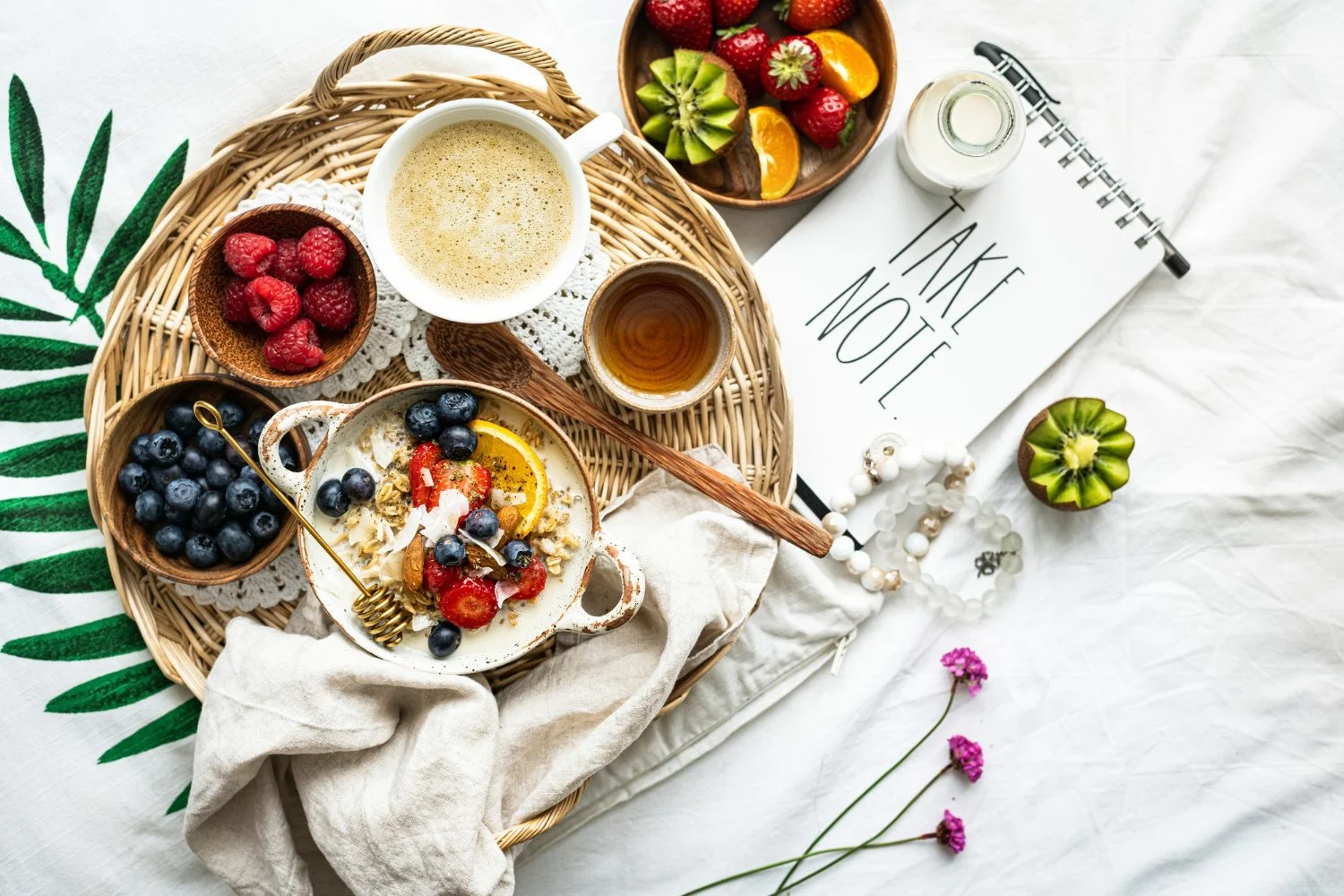 A breakfast tray with bowls of raspberries, blueberries, and sliced strawberries, a cup of coffee, a glass of tea, and a bowl of salad with fruit, on a white blanket with a 'Take Note!' notepad, jewelry, and pink flowers nearby.