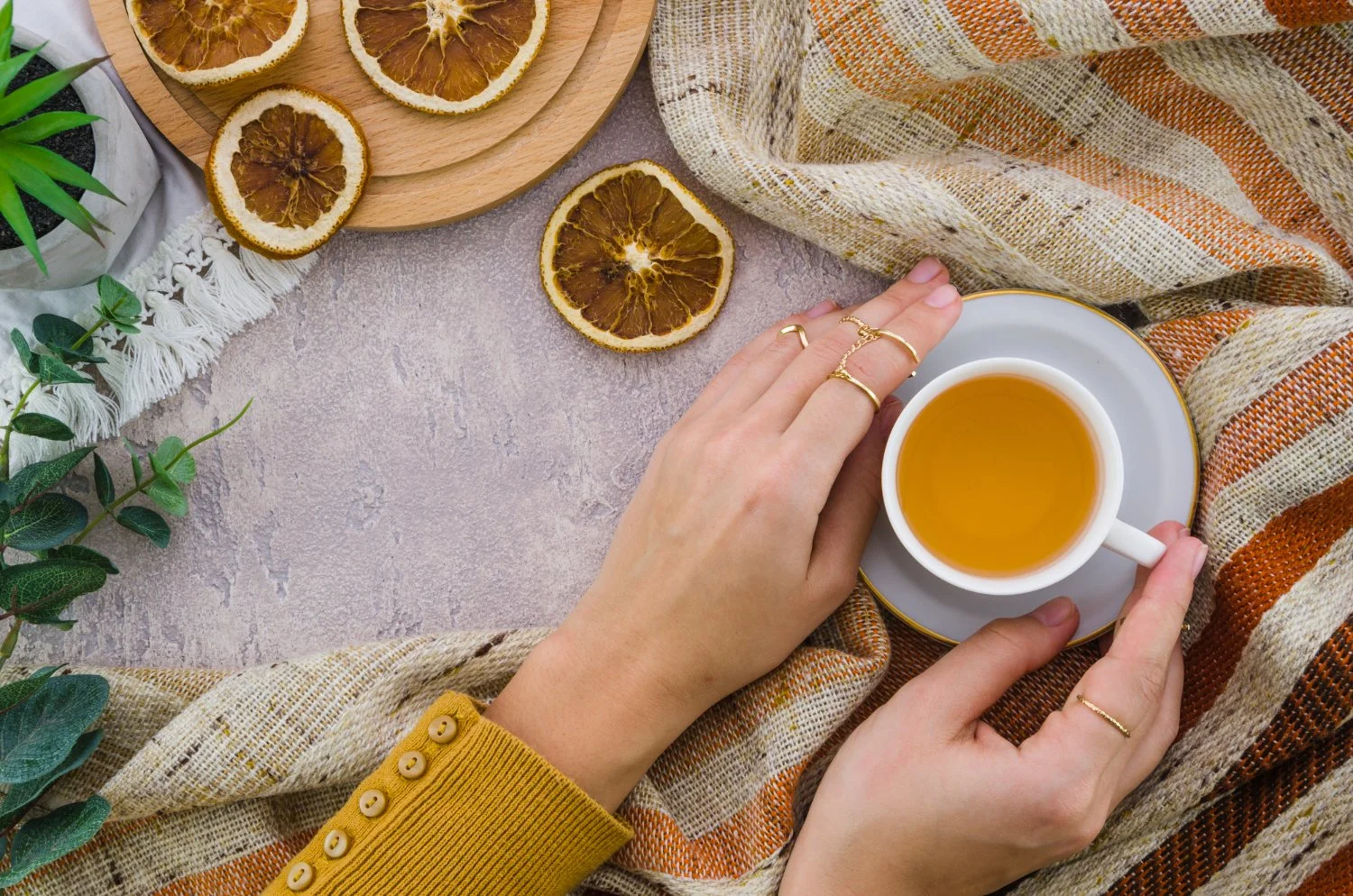 A person's hands holding a white teacup filled with tea, surrounded by dried lemon slices, a plaid scarf, plants, and a wooden tray on a textured surface.