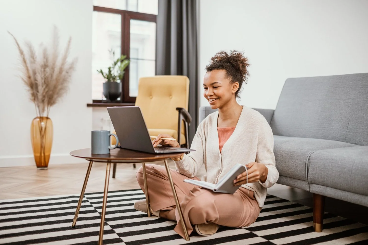 A woman sitting cross-legged on a striped rug, working on a laptop placed on a small round table, holding a notebook in one hand and smiling.