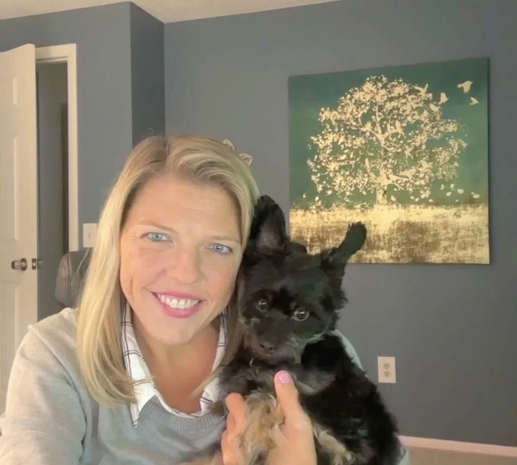 A woman with blonde hair smiling and holding a black puppy in front of a blue wall with a framed print of a tree.
