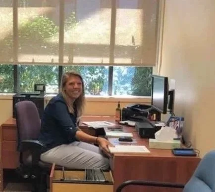 A woman sitting at a desk with a computer monitor, papers, and office supplies, in an office with windows showing greenery outside.