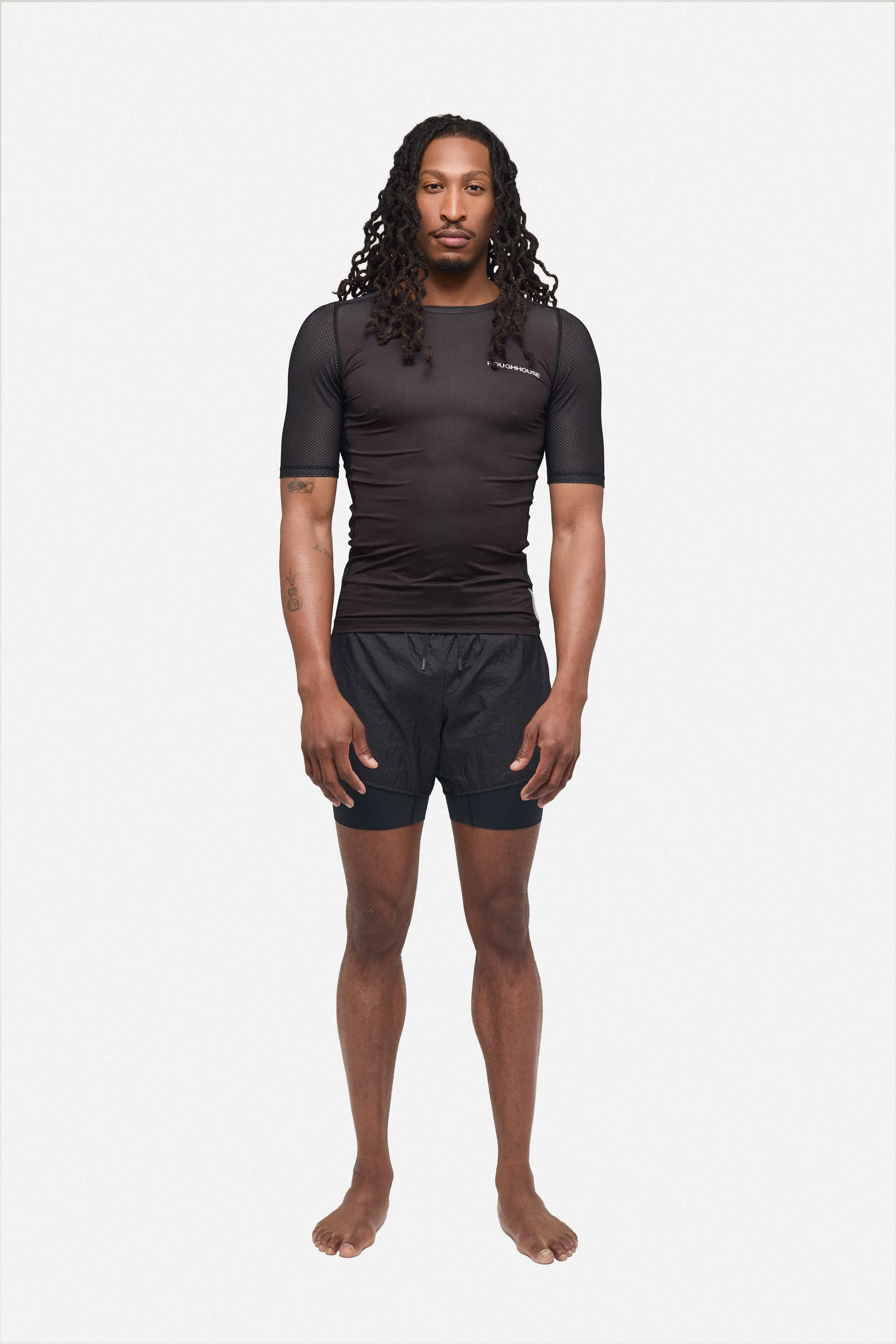 A man with long dreadlocks wearing a black athletic shirt and shorts, standing barefoot against a plain background.