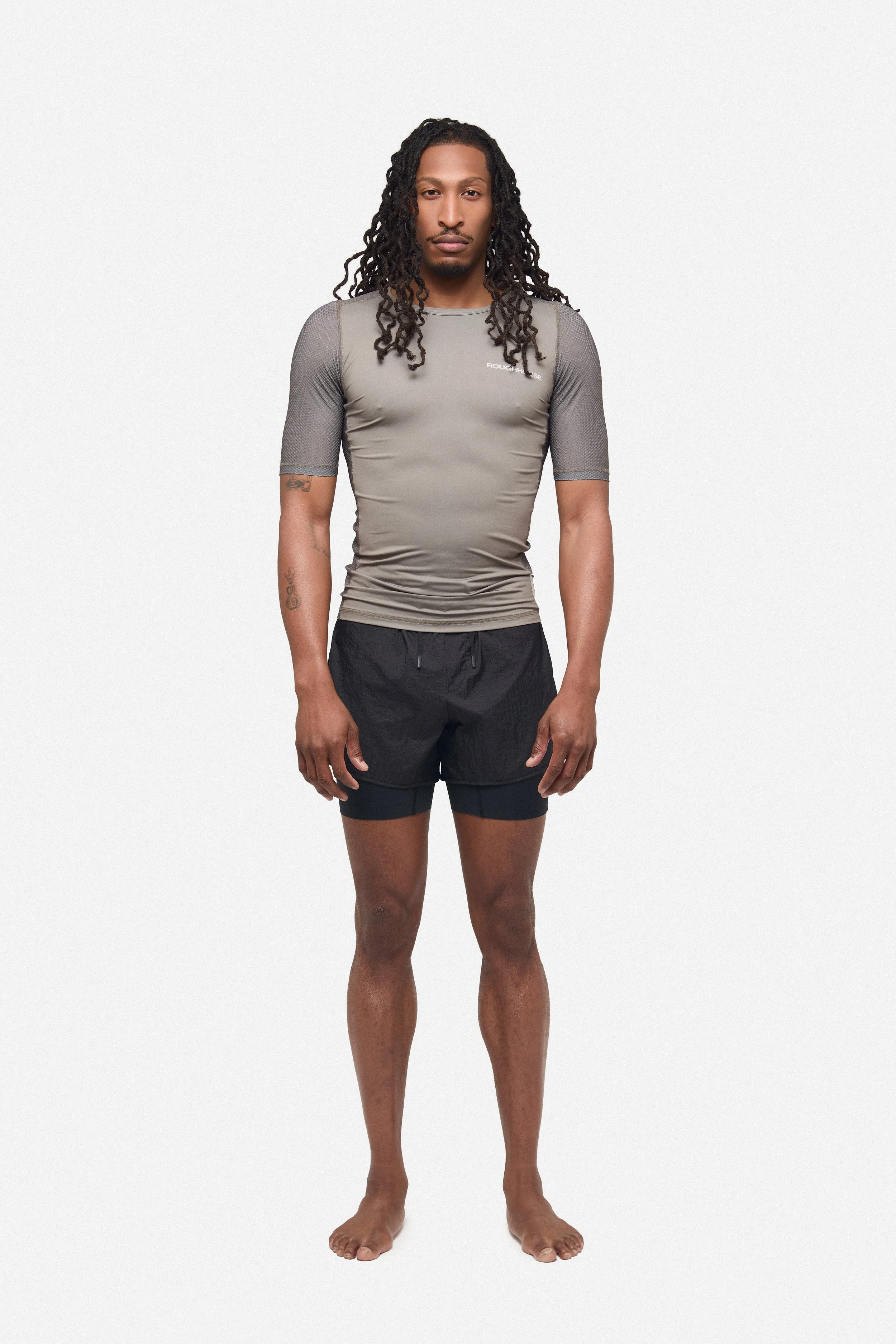A man standing barefoot against a plain white background, wearing a fitted gray athletic shirt with mesh sleeves and black shorts.