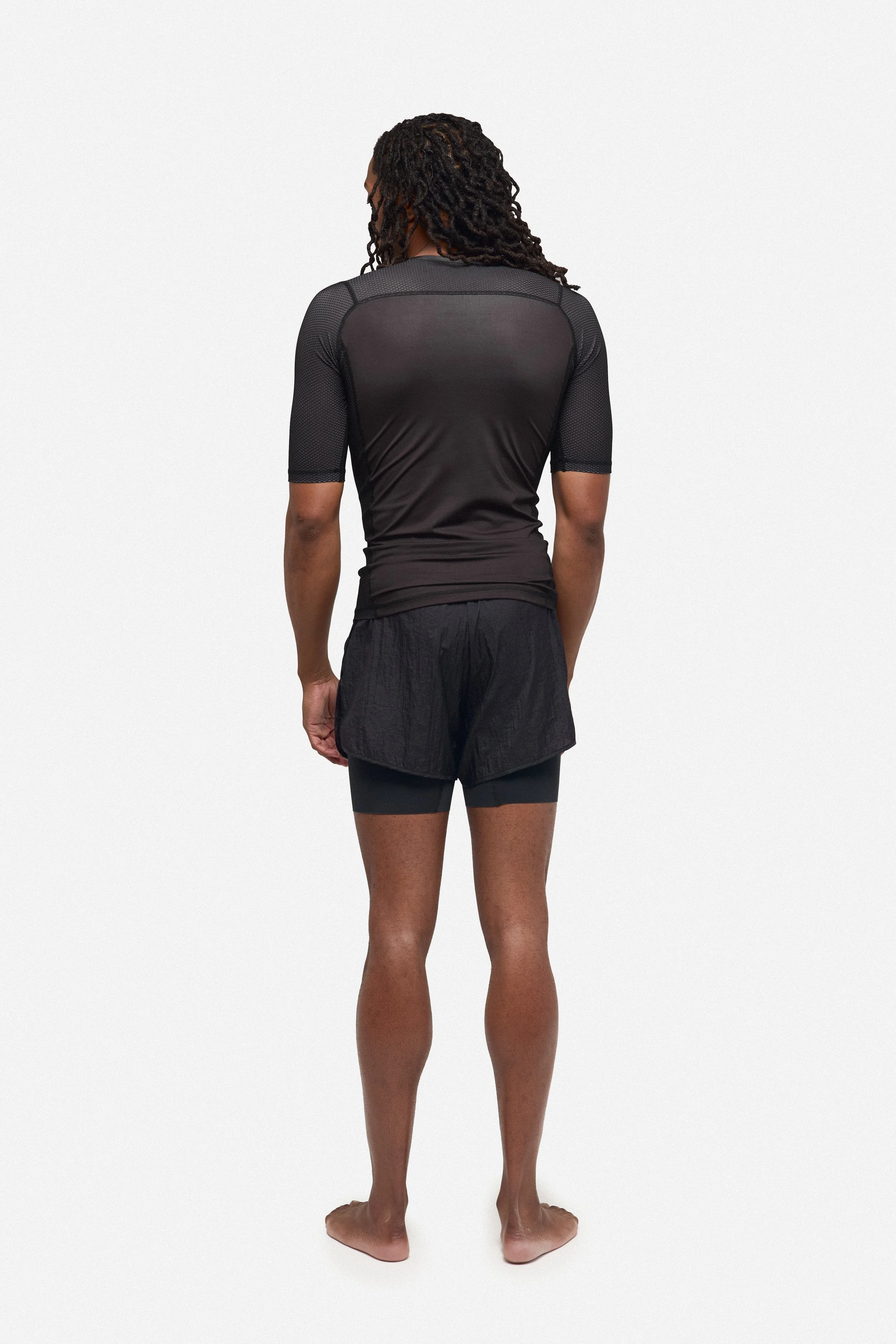 Person standing barefoot, wearing black athletic shorts and a black short-sleeved, mesh athletic shirt, facing away from the camera against a plain white background.