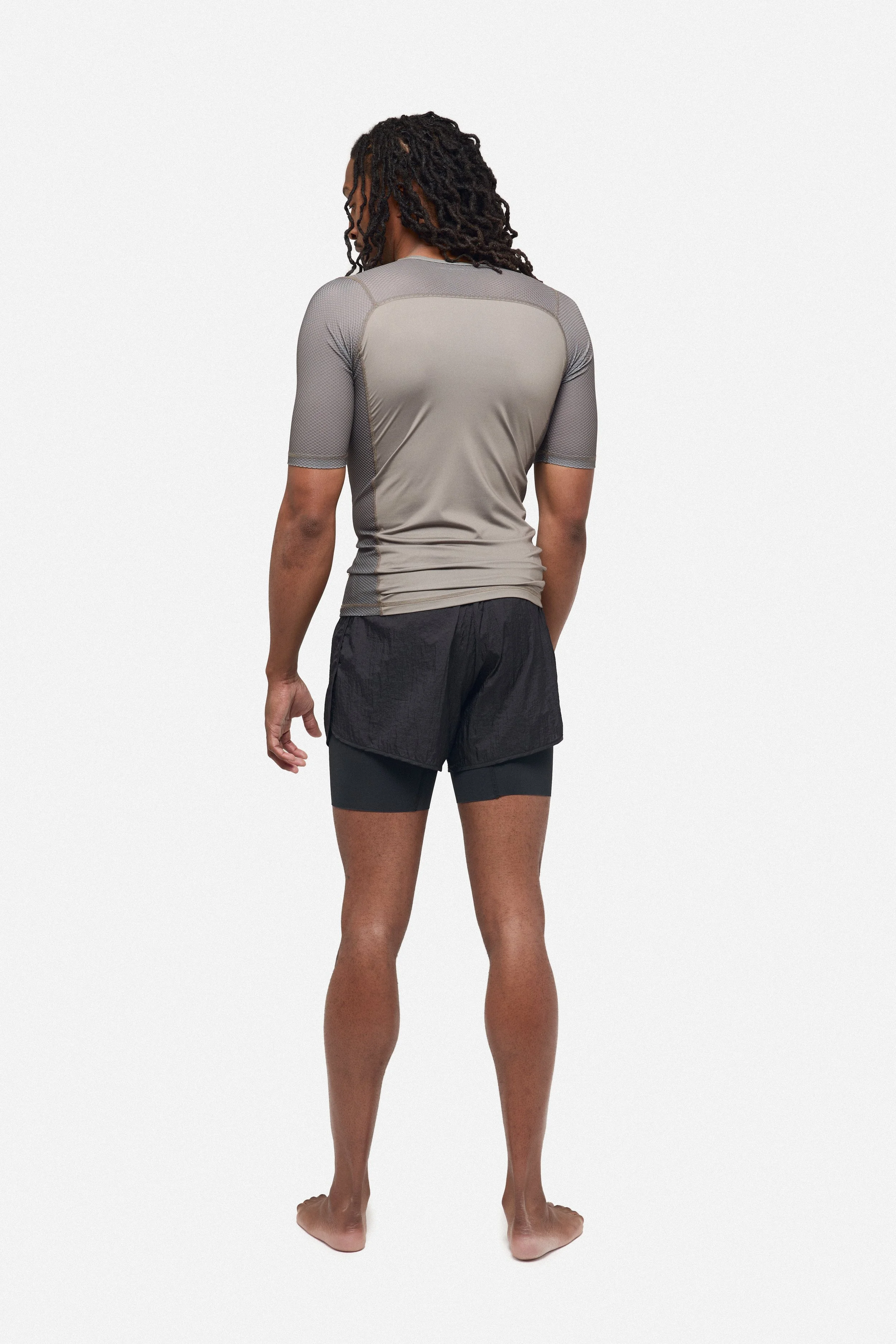 Back view of a man with dreadlocks wearing a gray athletic shirt, black shorts, and standing barefoot against a plain white background.
