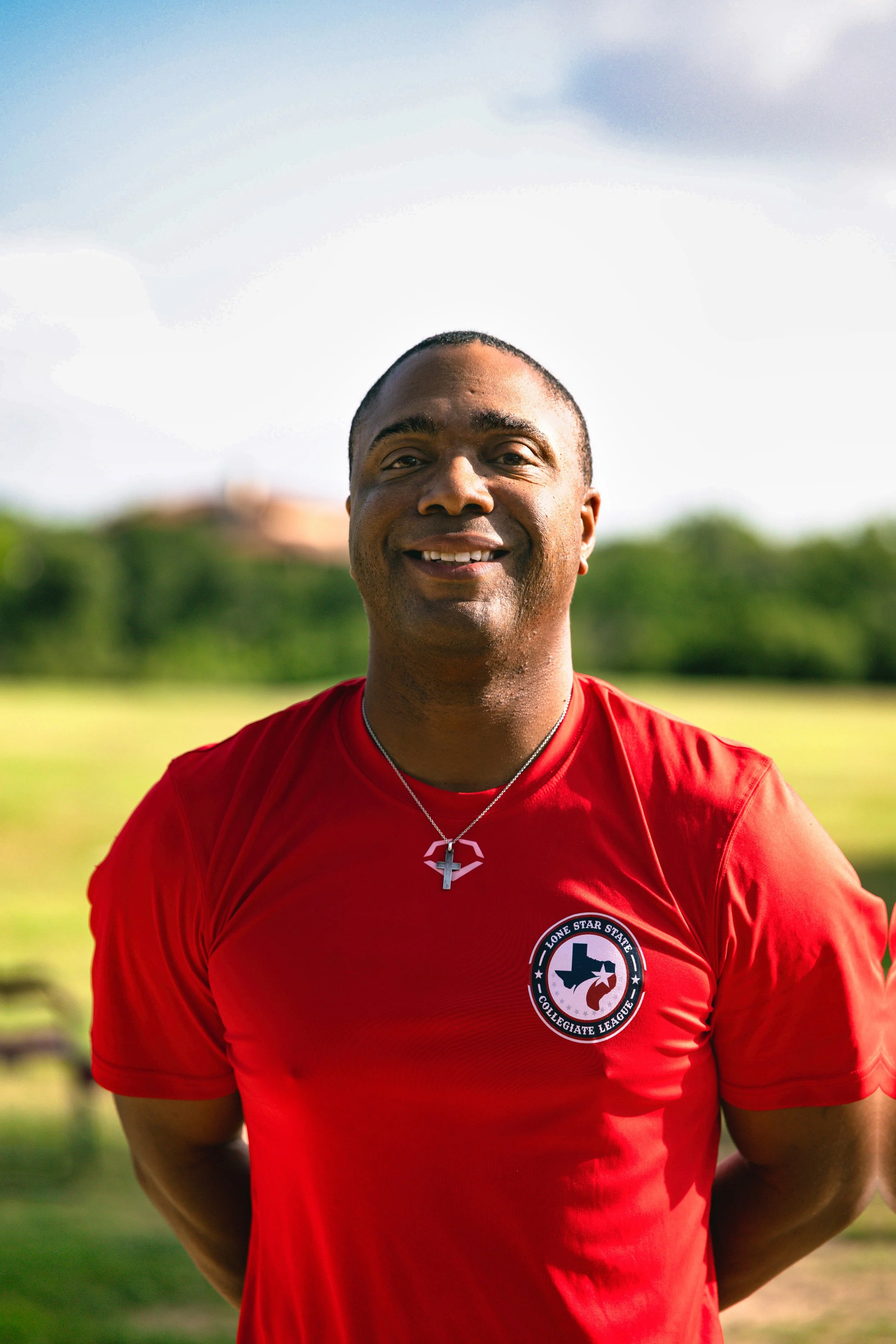 A man wearing a red sports shirt with a Lone Star State Collegiate League logo, standing outdoors on a bright, sunny day. He is smiling and wearing a cross necklace, with a blurred green field and trees in the background.