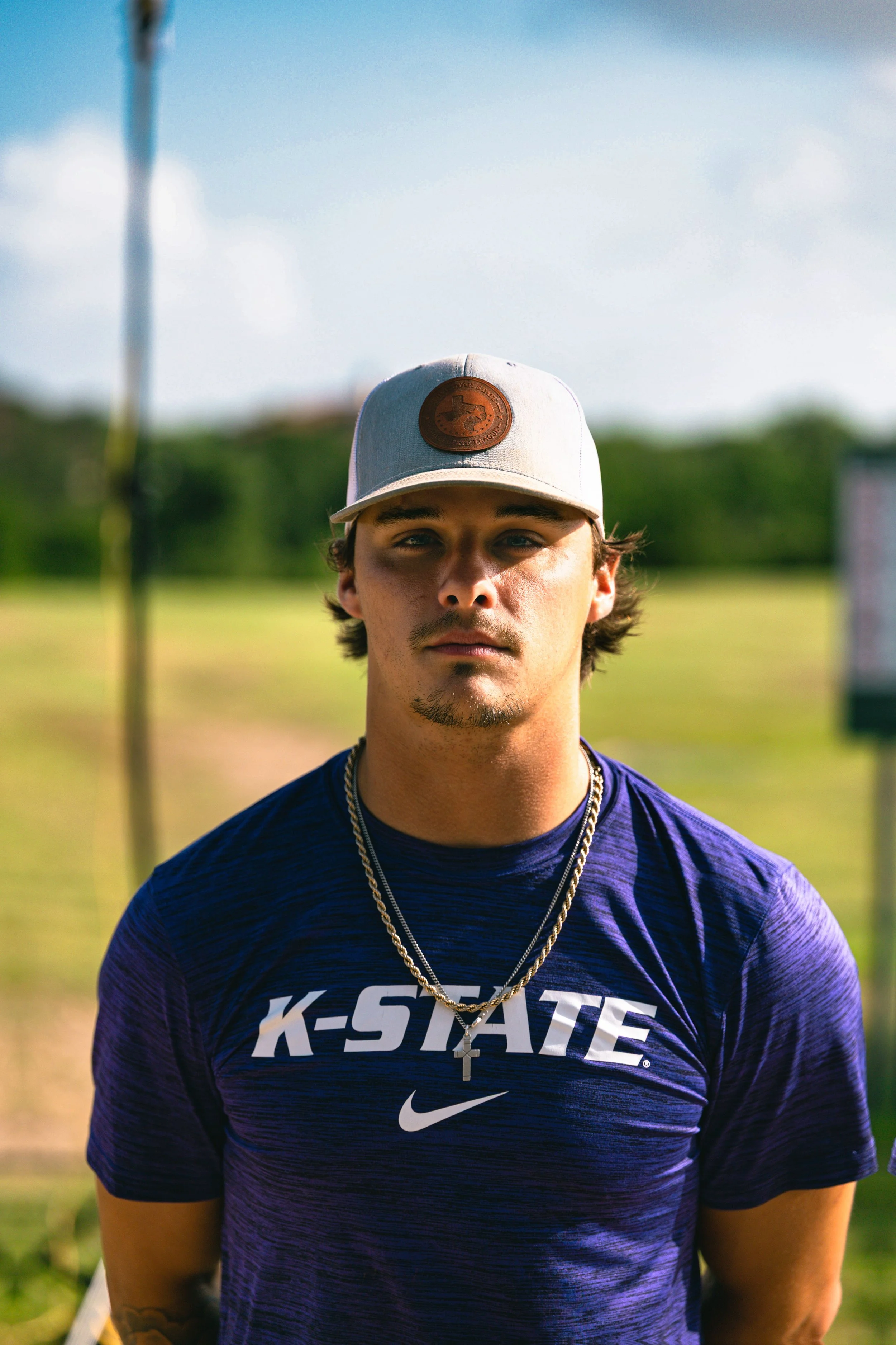 Young man wearing a gray baseball cap with a brown logo, a purple K-State shirt, and gold necklaces, standing outdoors with a blurred green background and blue sky.