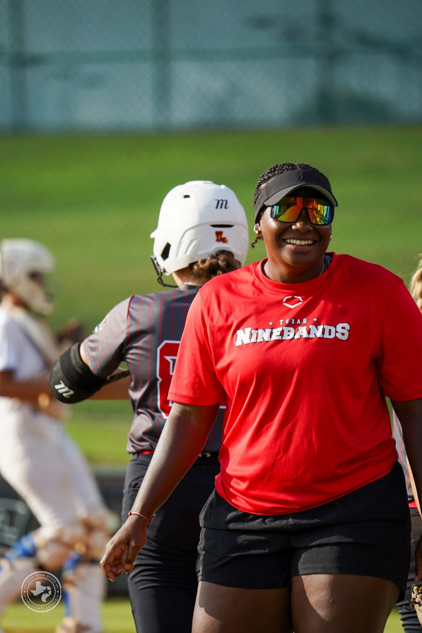 A woman sitting on a red YETI cooler on a baseball field, writing in a notebook, wearing red athletic shoes, a red T-shirt, black shorts, sunglasses, a visor, and a wristwatch, with a football field and trees in the background.