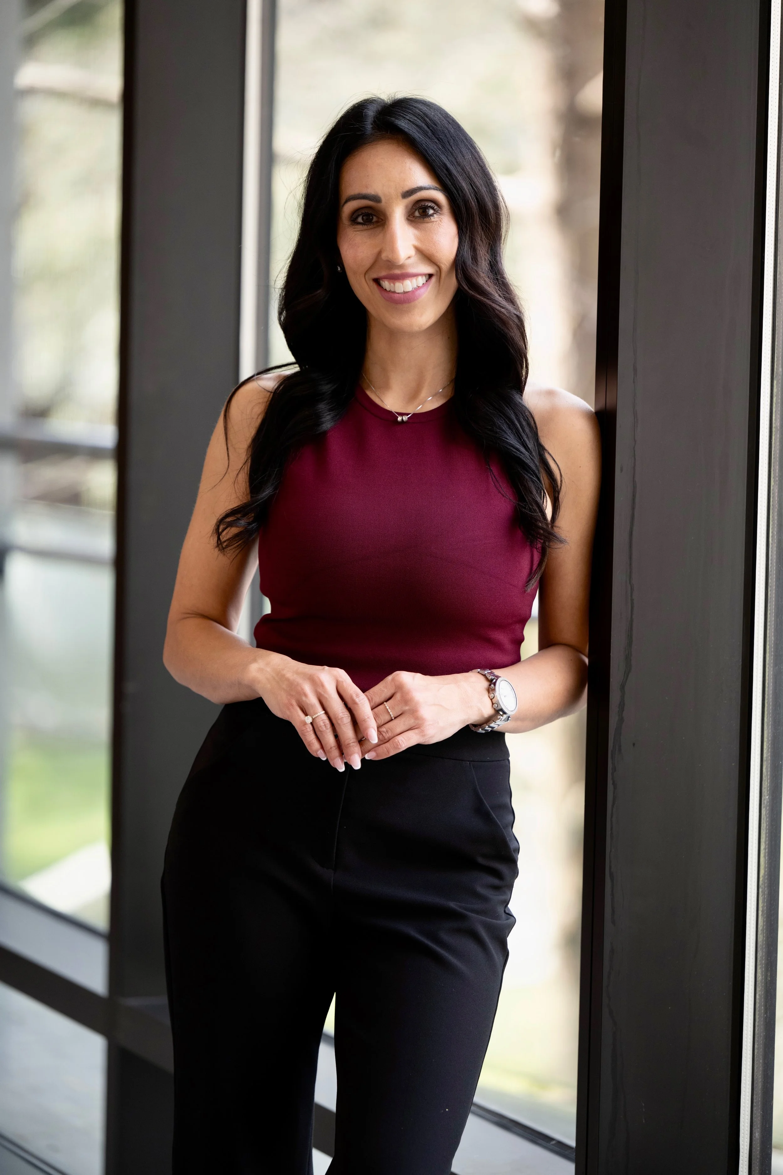 A woman with long black hair, wearing a sleeveless maroon top and black pants, standing by a large window with a blurred outdoor background, smiling at the camera.