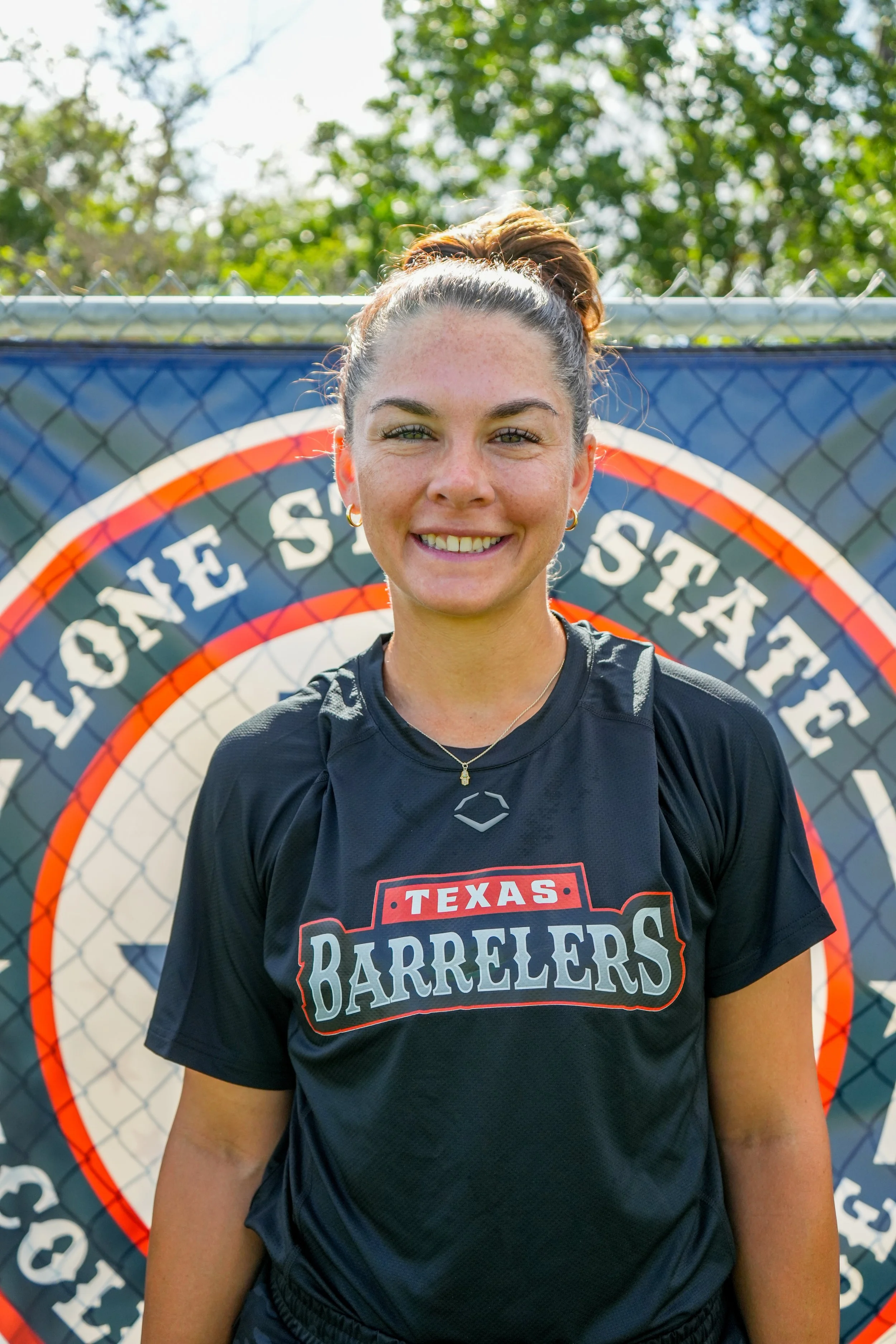 A young woman with brown hair in a ponytail, smiling, wearing a black sports jersey with 'Texas Barrelers' written on it, standing outdoors in front of a fence with a sign that says 'Lone Star'.