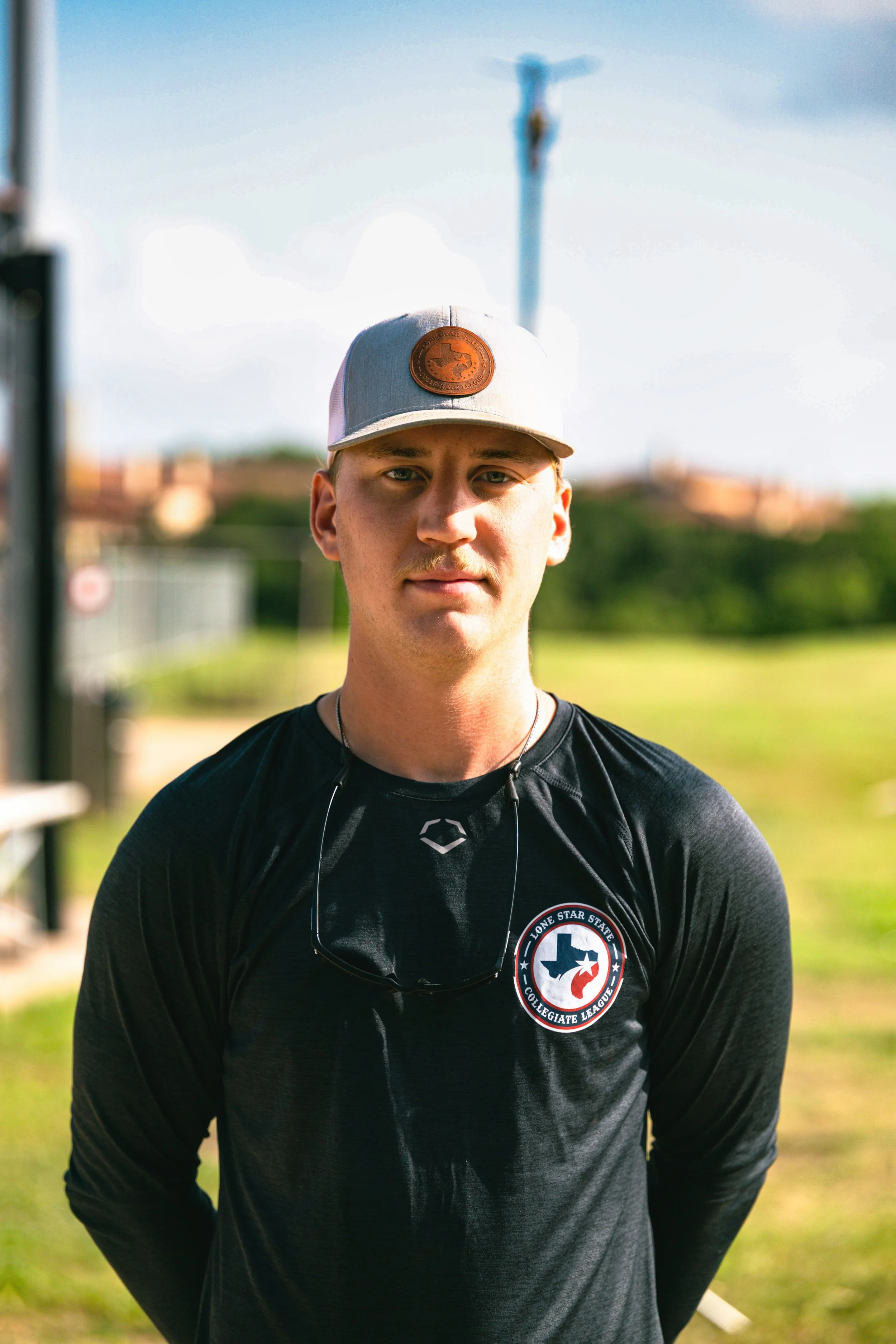 Young man outdoors wearing a black sports shirt with a Texas flag and baseball emblem, a gray cap, and sunglasses hanging around his neck, standing in front of a sports field with a baseball goal and a flagpole