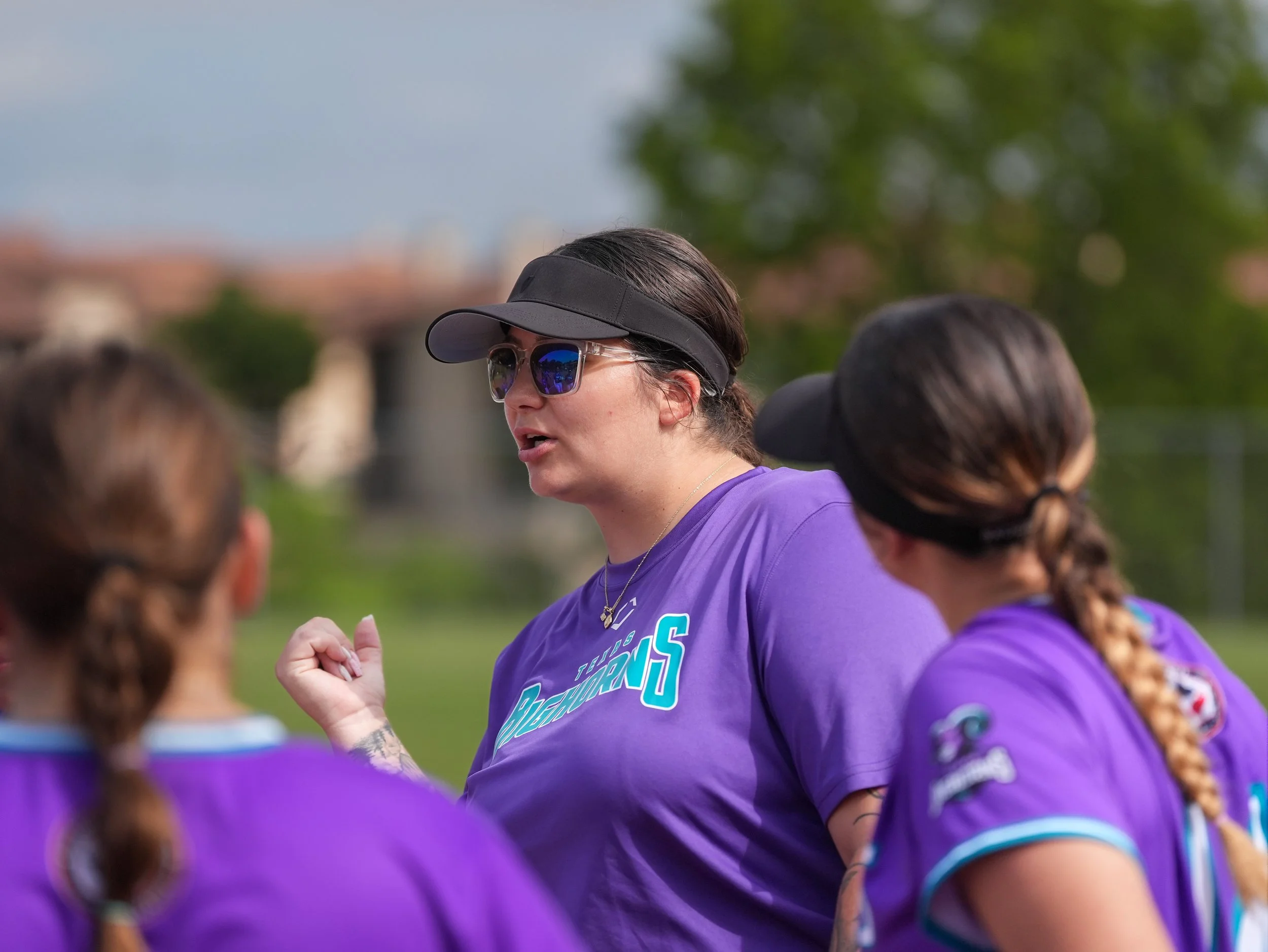 A female softball coach with sunglasses and a black visor speaks to players in purple jerseys during a game.