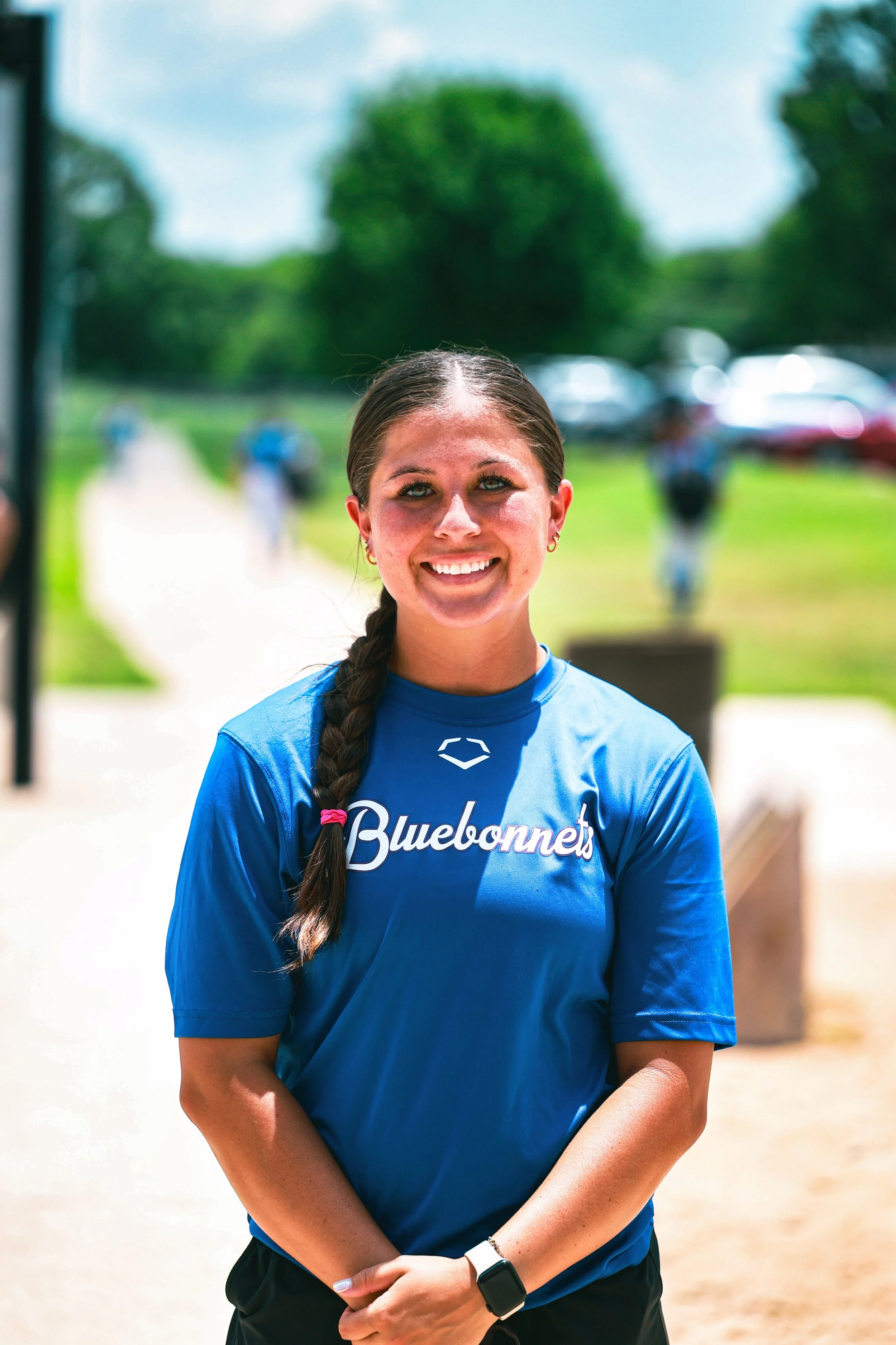 A woman with long braided hair wearing a blue 'Bluebonnets' t-shirt, smiling outdoors on a sunny day.