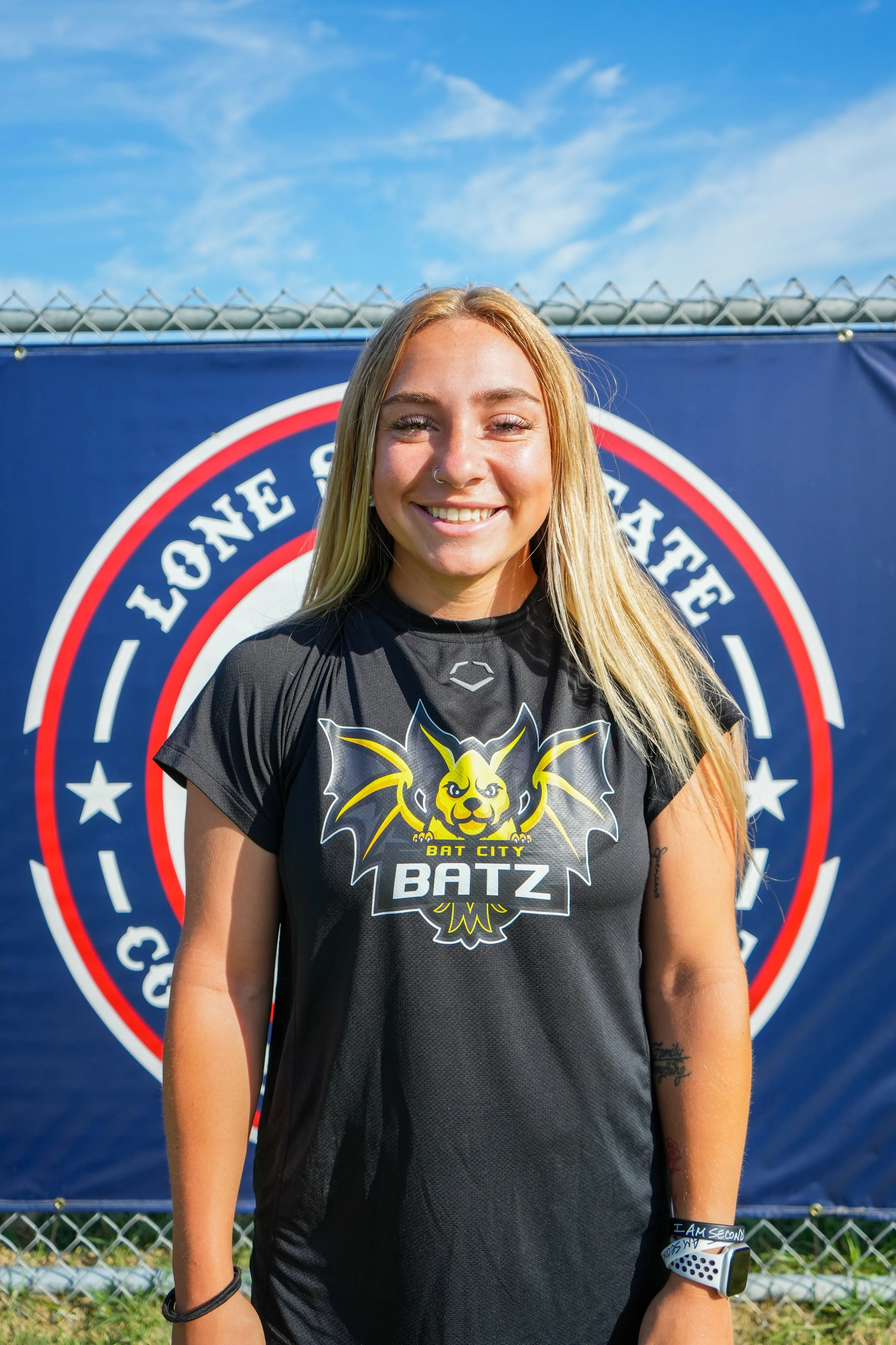 Young woman with long blonde hair and a nose ring smiling, wearing a black Bat City BATZ jersey, standing outdoors in front of a large circular team logo banner, with a chain-link fence and a partly cloudy blue sky in the background.