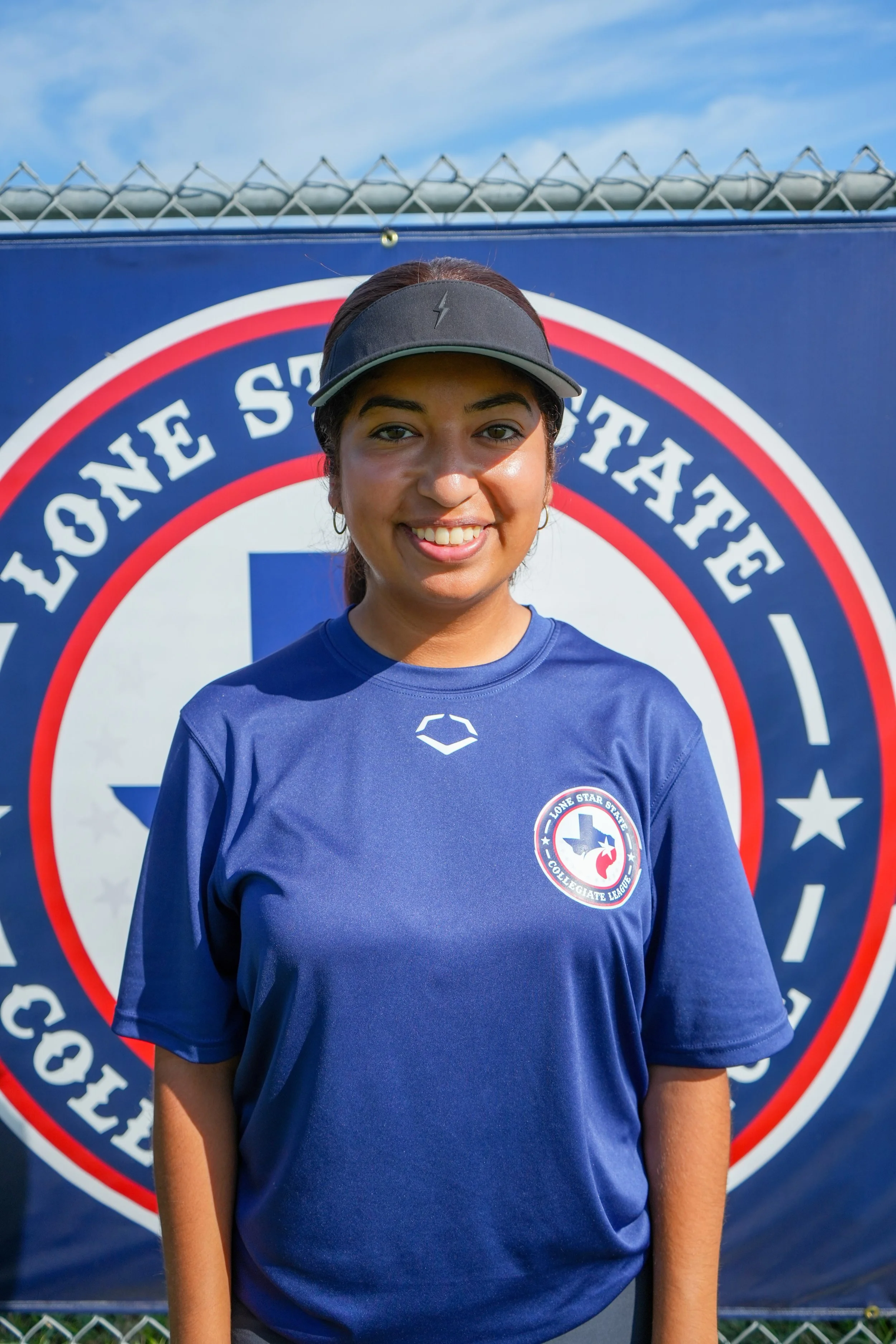 A young woman smiling in front of a Lone Star State Collegiate League banner, wearing a blue T-shirt and black visor.