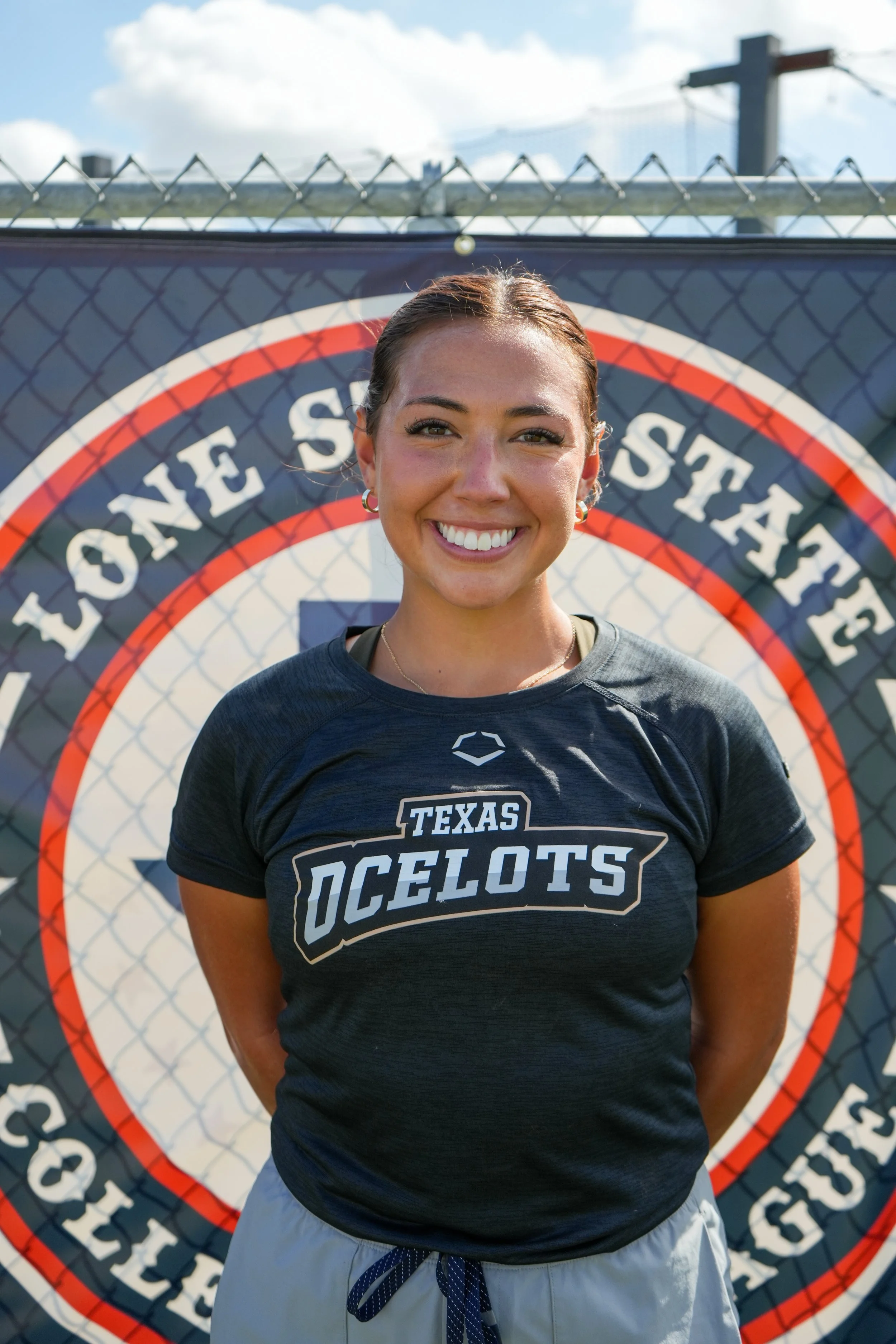 Smiling woman in athletic wear standing outdoors in front of a fence with a large Texas Longstone State soccer logo.