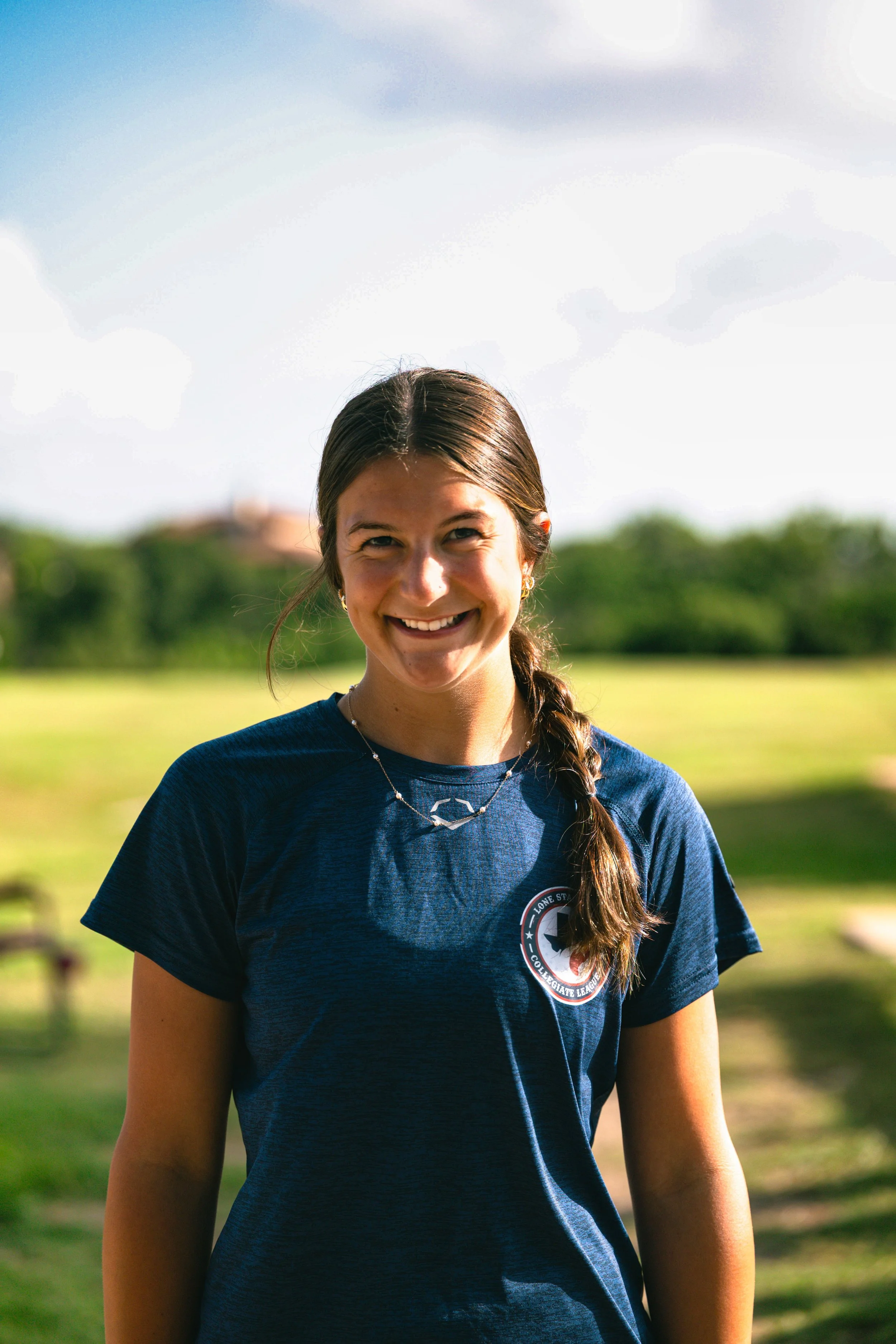 A young woman smiling outdoors in a park on a sunny day, wearing a navy blue athletic shirt with a logo, and a necklace with a star-shaped pendant.