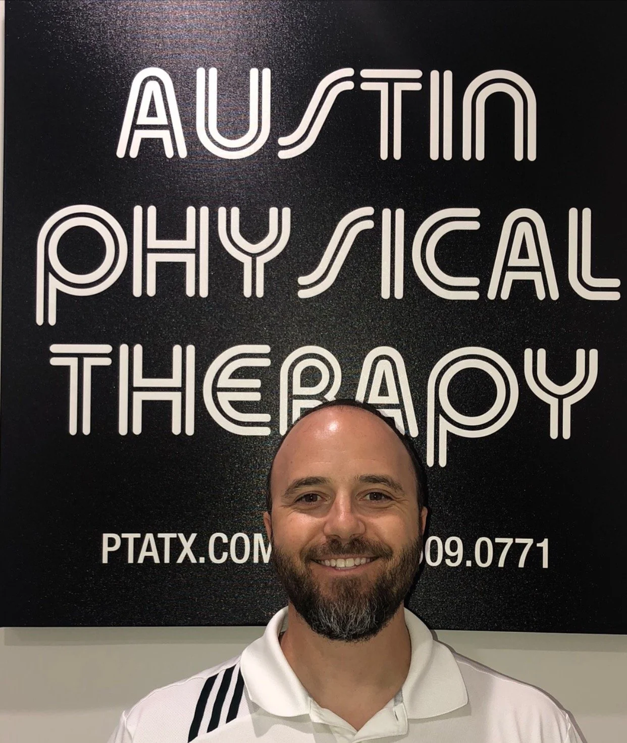 Man smiling in front of a black sign reading "AUSTIN PHYSICAL THERAPY" with website and phone number.