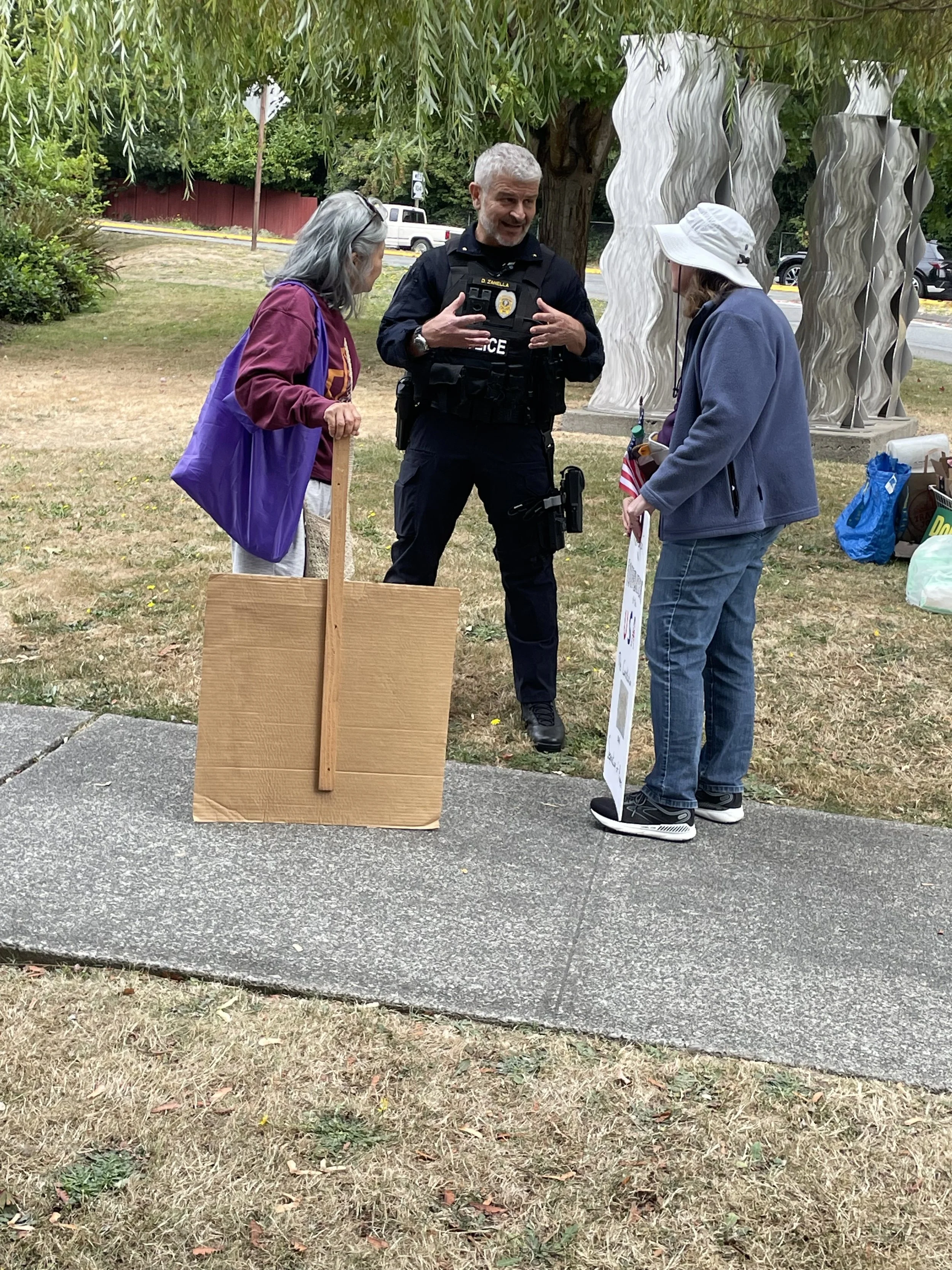 A police officer engaged in conversation with two women outdoors, with a wooden protest sign and a large poster board.