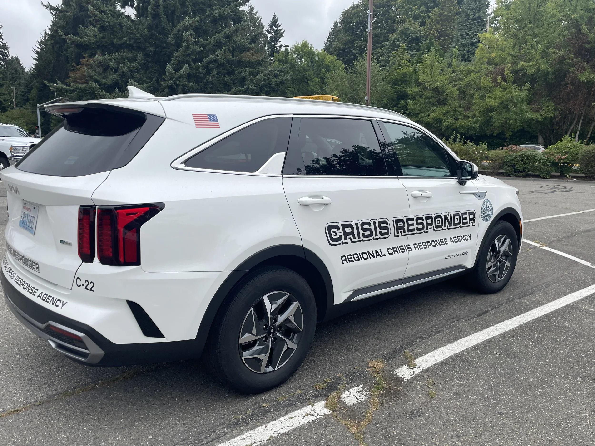 A white crisis response vehicle parked in a parking lot with trees in the background. The vehicle has black lettering that reads 'CRISIS RESPONDER' and 'REGIONAL CRISIS RESPONSE AGENCY.' It has a small American flag decal on the rear side window and a license plate in the front.