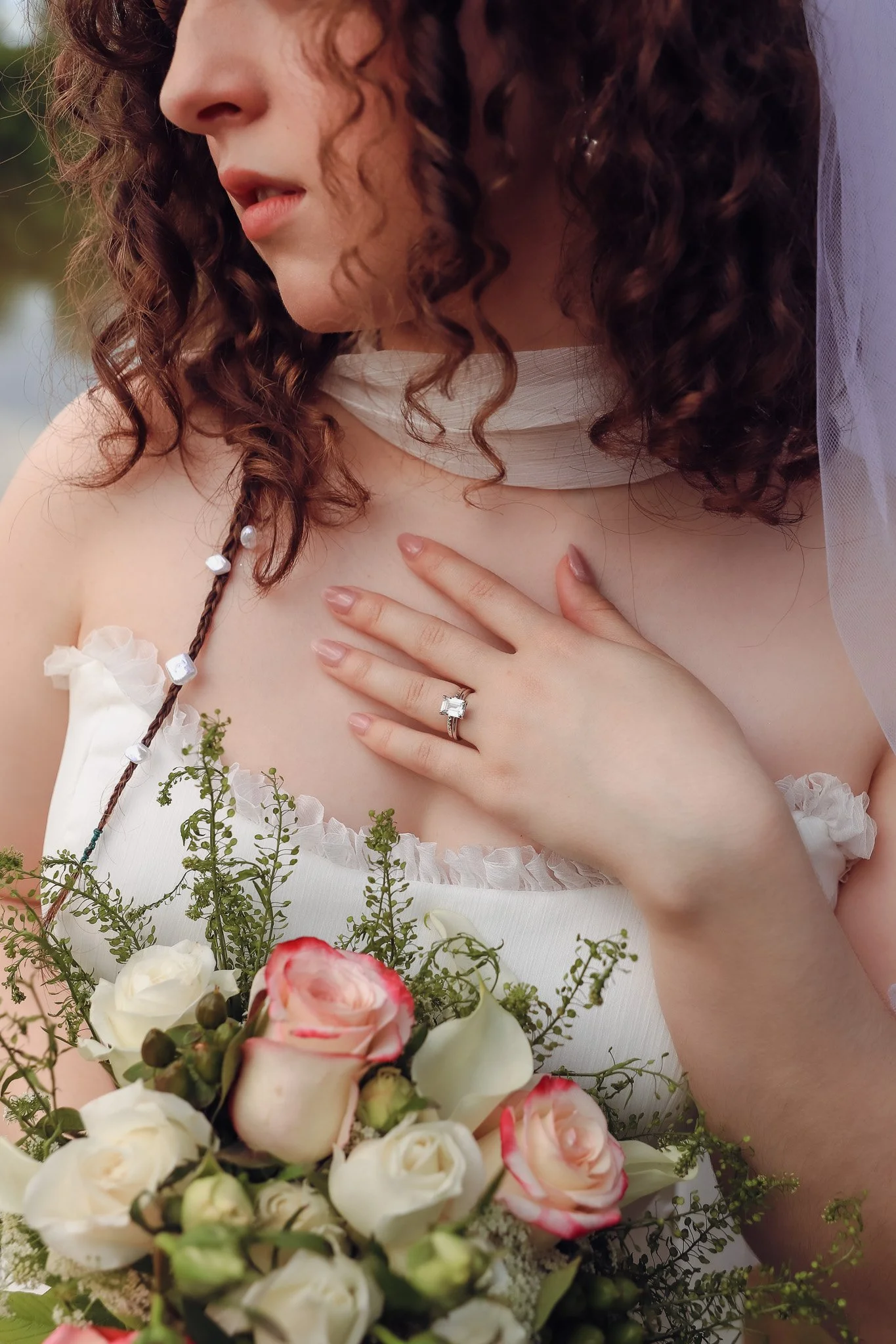 A bride wearing a wedding dress and veil, holding a bouquet of white and pink roses, showing her hand with a diamond ring on her finger, with curly hair and a soft expression.